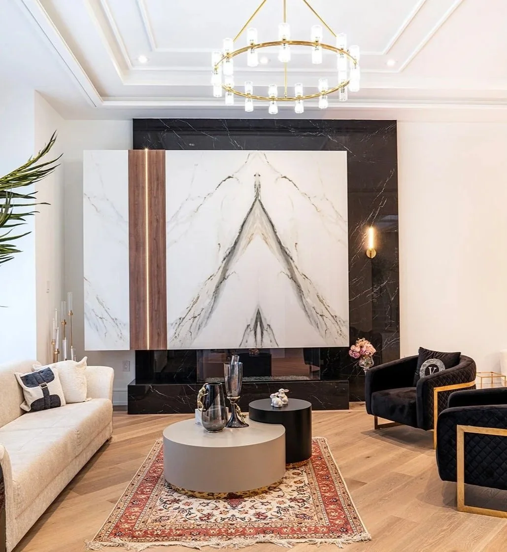Living room with modern decor, large marble fireplace with black and white marble, a beige sofa with pillows, two black velvet chairs with gold frames, a gold chandelier, round coffee tables, and a patterned rug.