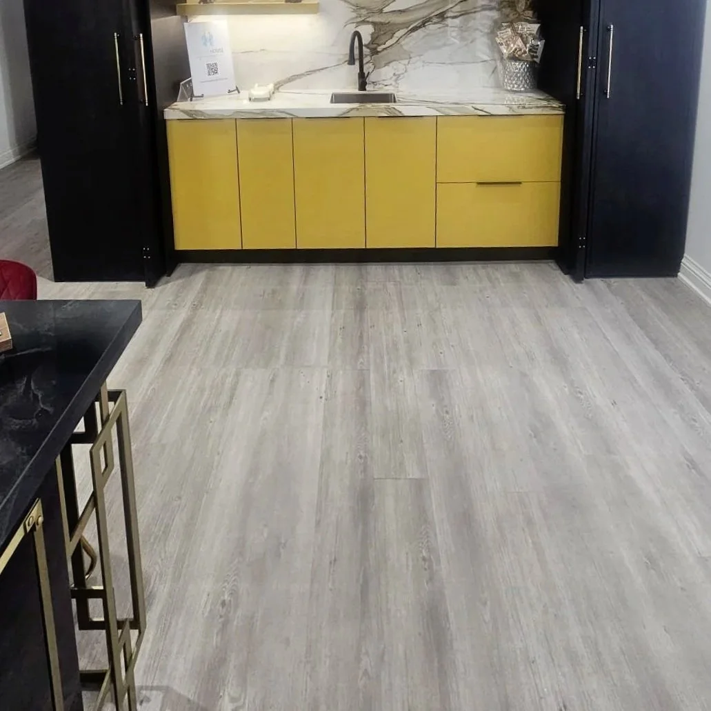 A kitchen with countertop, yellow cabinet base, black upper cabinets, and a marble backsplash. There is a sink and faucet beneath the backsplash, with a small sign and a tissue holder nearby. The floor is a light gray wood texture.