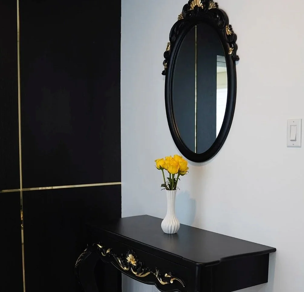 A black ornate wall mirror with gold accents hangs above a black console table with gold accents. A white vase with yellow flowers sits on the table, against a white wall with a light switch nearby.