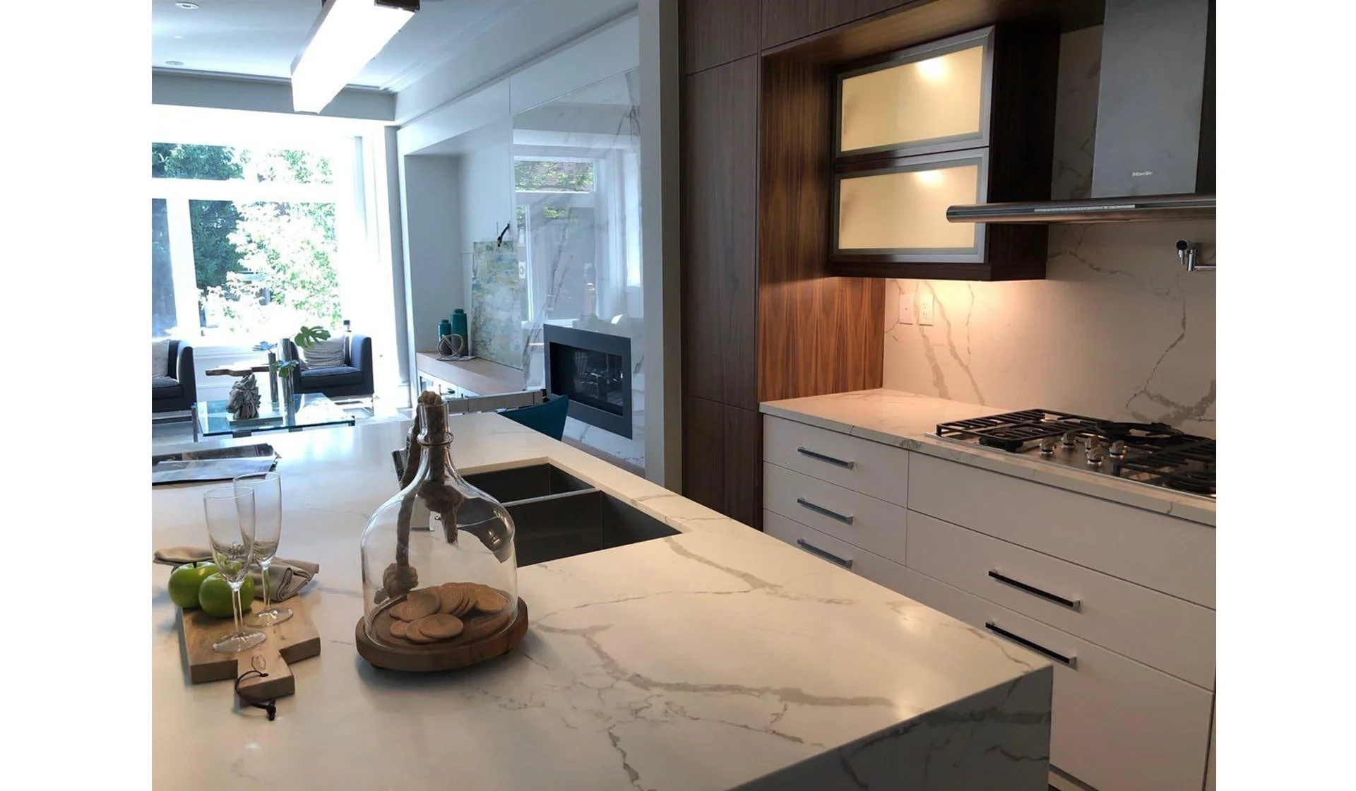 Modern kitchen with white marble countertops, wooden cabinets, a built-in gas stove, a sink, and decorative items on the island, with a seating area and large windows in the background.