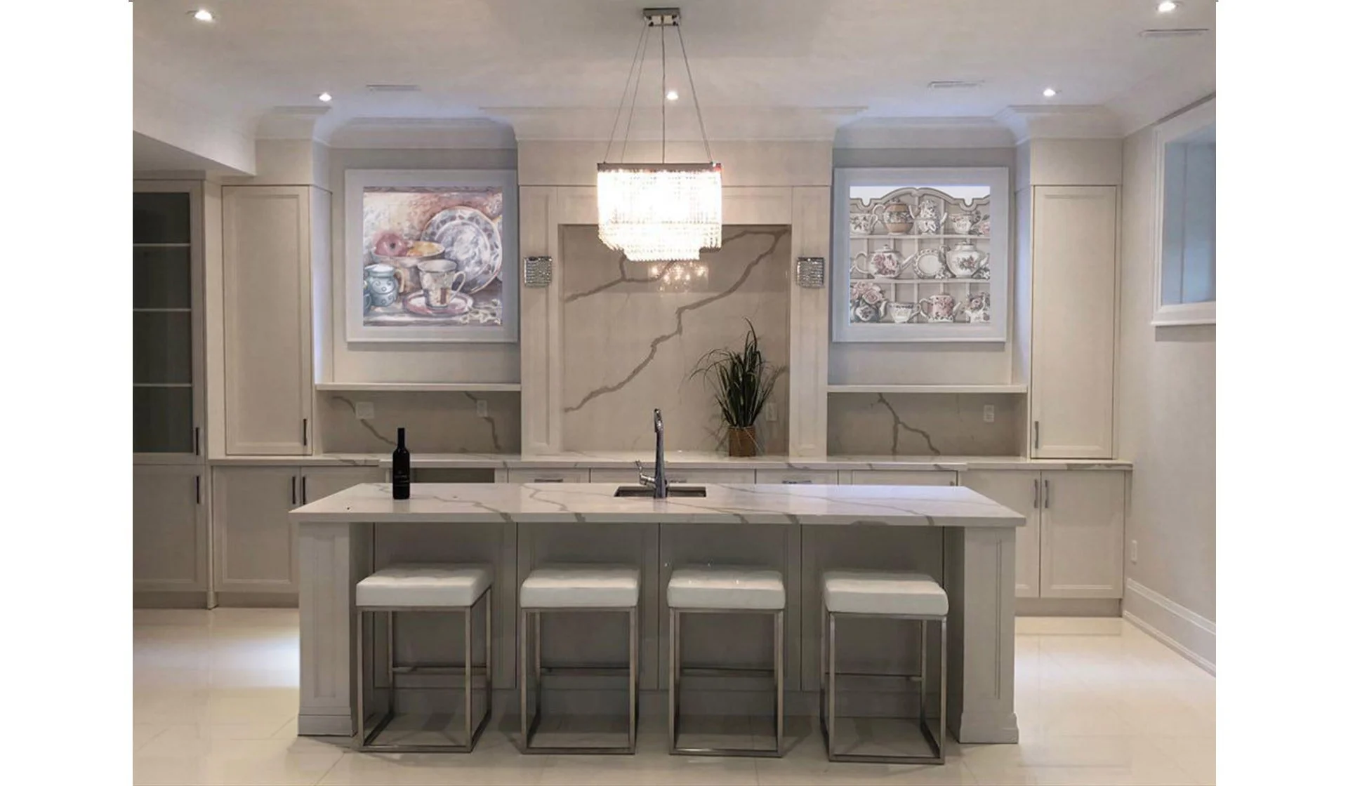 Modern kitchen with a large island, white cabinetry, marble countertops, and a chandelier. Two built-in display shelves with decorative dishware on the walls behind the island.