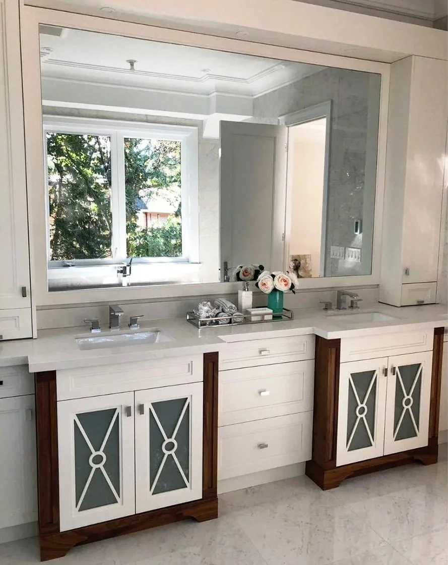 Bathroom double vanity with white cabinets, glass-paneled doors, and a marble countertop. A large mirror spans the wall above, reflecting a window with greenery outside. Decor includes a green vase with flowers and toiletries.