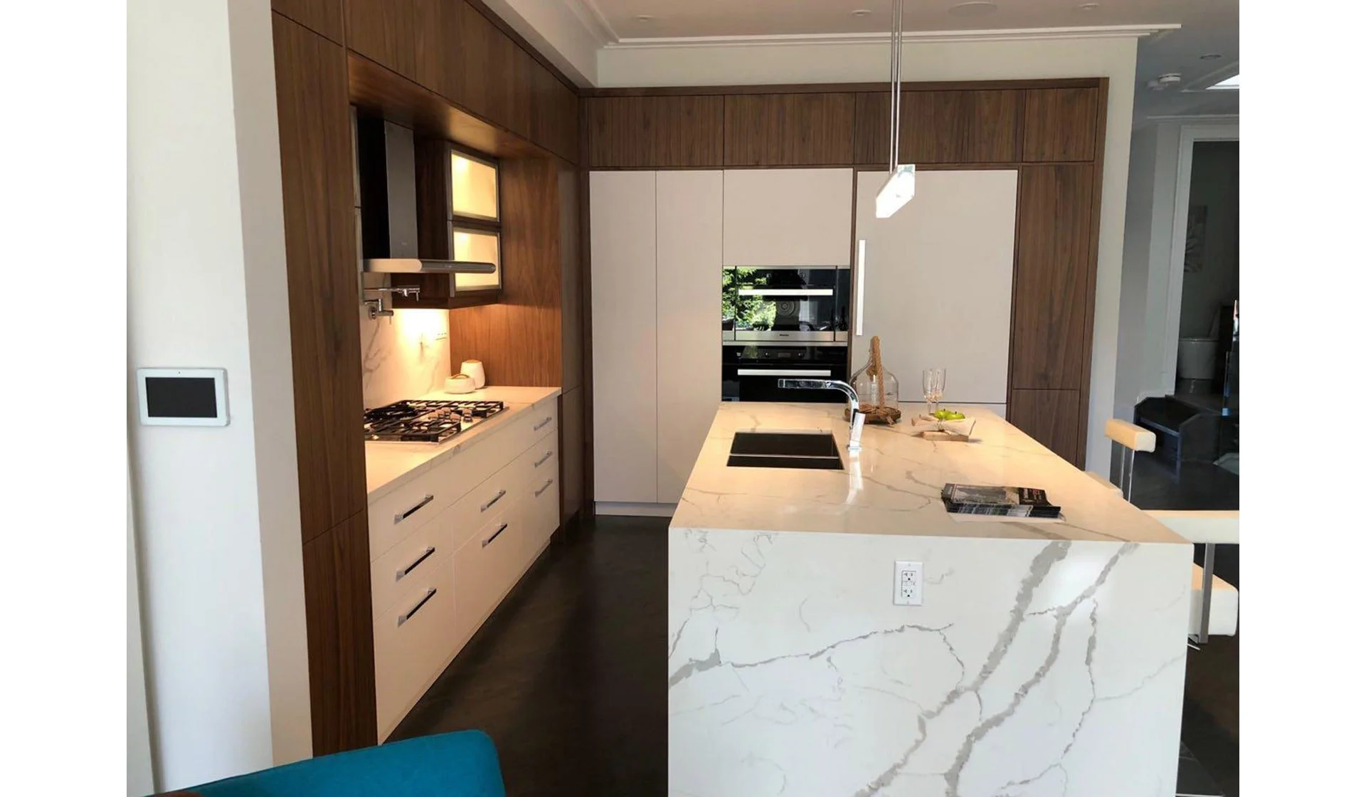 Modern kitchen with white marble island, wooden wall cabinets, and built-in appliances. The kitchen has a minimalist design with a single hanging light and a view into another room.