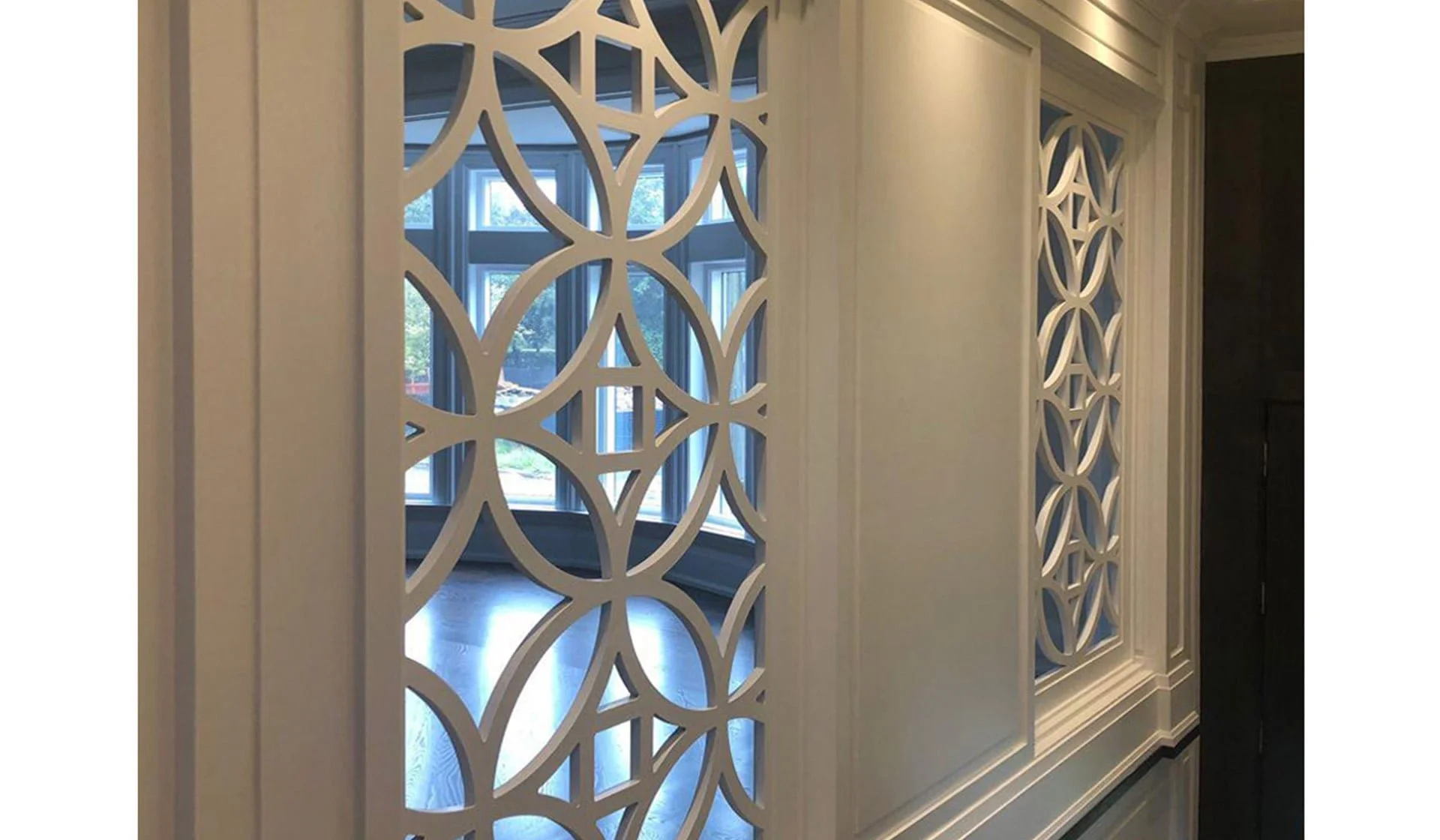Decorative white lattice panels with geometric patterns, mounted on a wall in a well-lit interior space with large windows showing outdoor greenery.