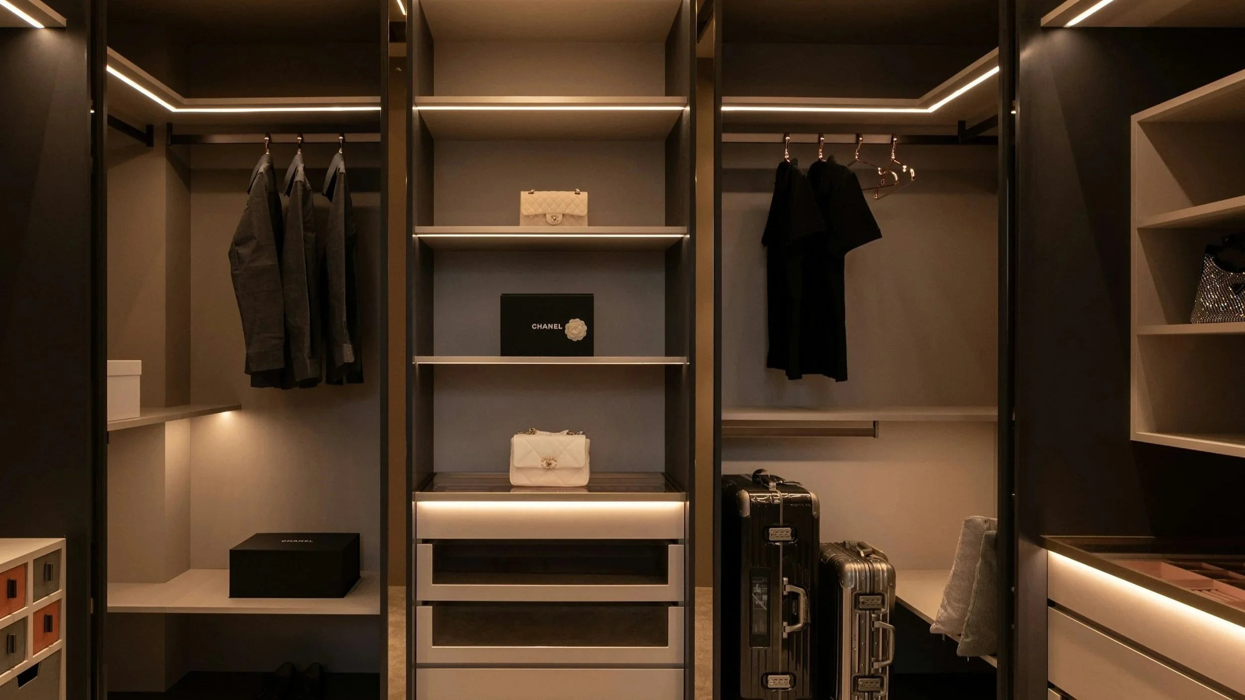 Luxury walk-in closet with hanging clothes, handbags, and luggage, featuring modern lighting and organized shelves.