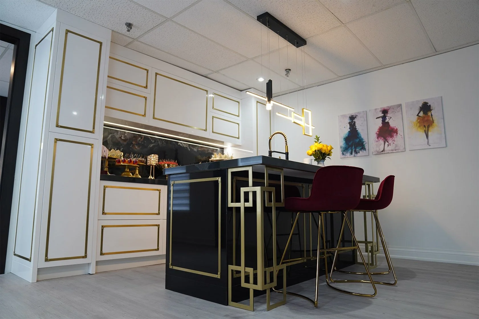 Modern kitchen with black island featuring gold geometric accents, white cabinets with gold trim, and marbled backsplash. There are two burgundy chairs with gold legs, a vase of yellow flowers, and colorful abstract art on the wall.