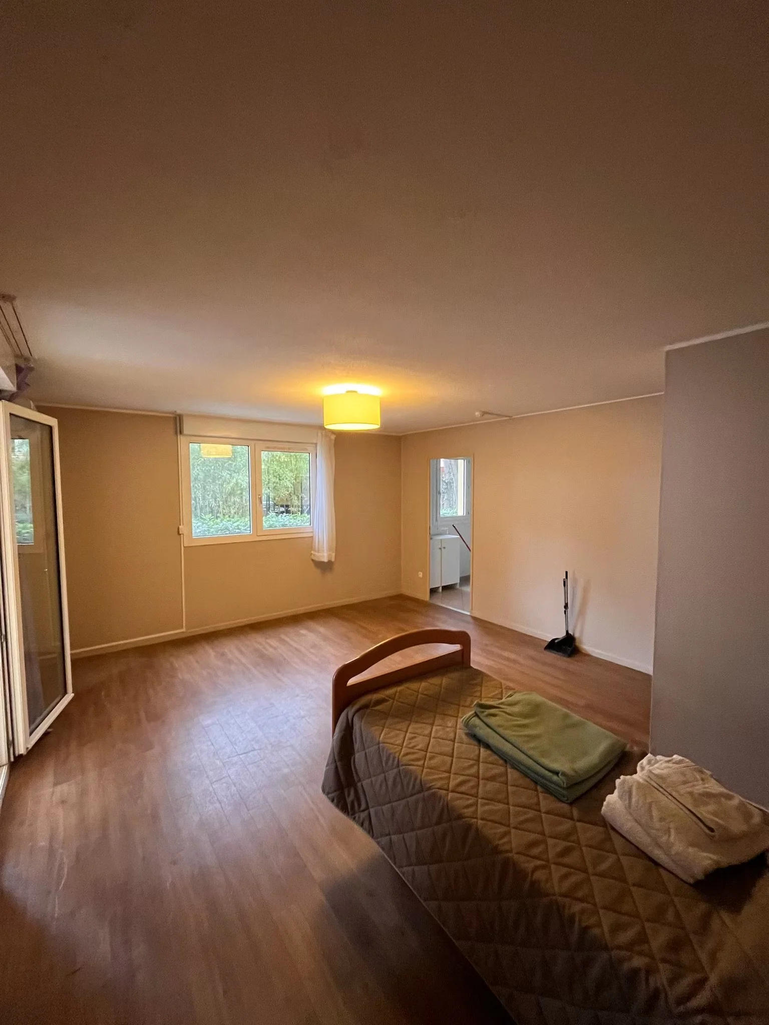 Chambre vide avec un lit, un tapis de bain, un aspirateur et des fenêtres laissant entrer la lumière naturelle.