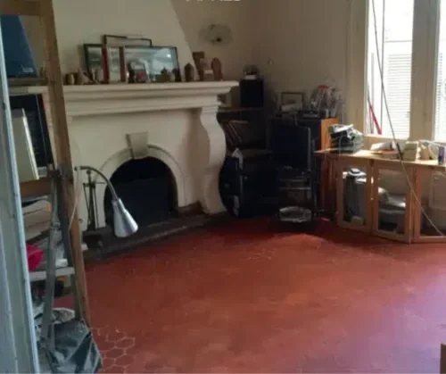 Salon avec cheminée blanche, bureau, et étagère à gauche, sol en carrelage rouge.