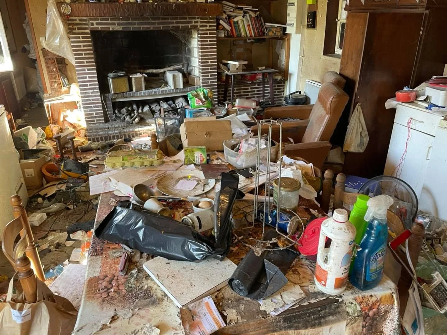 Une pièce en désordre avec une cheminée en briques, un mobilier en bois, une table couverte de déchets, objets divers, et des meubles encombrés.