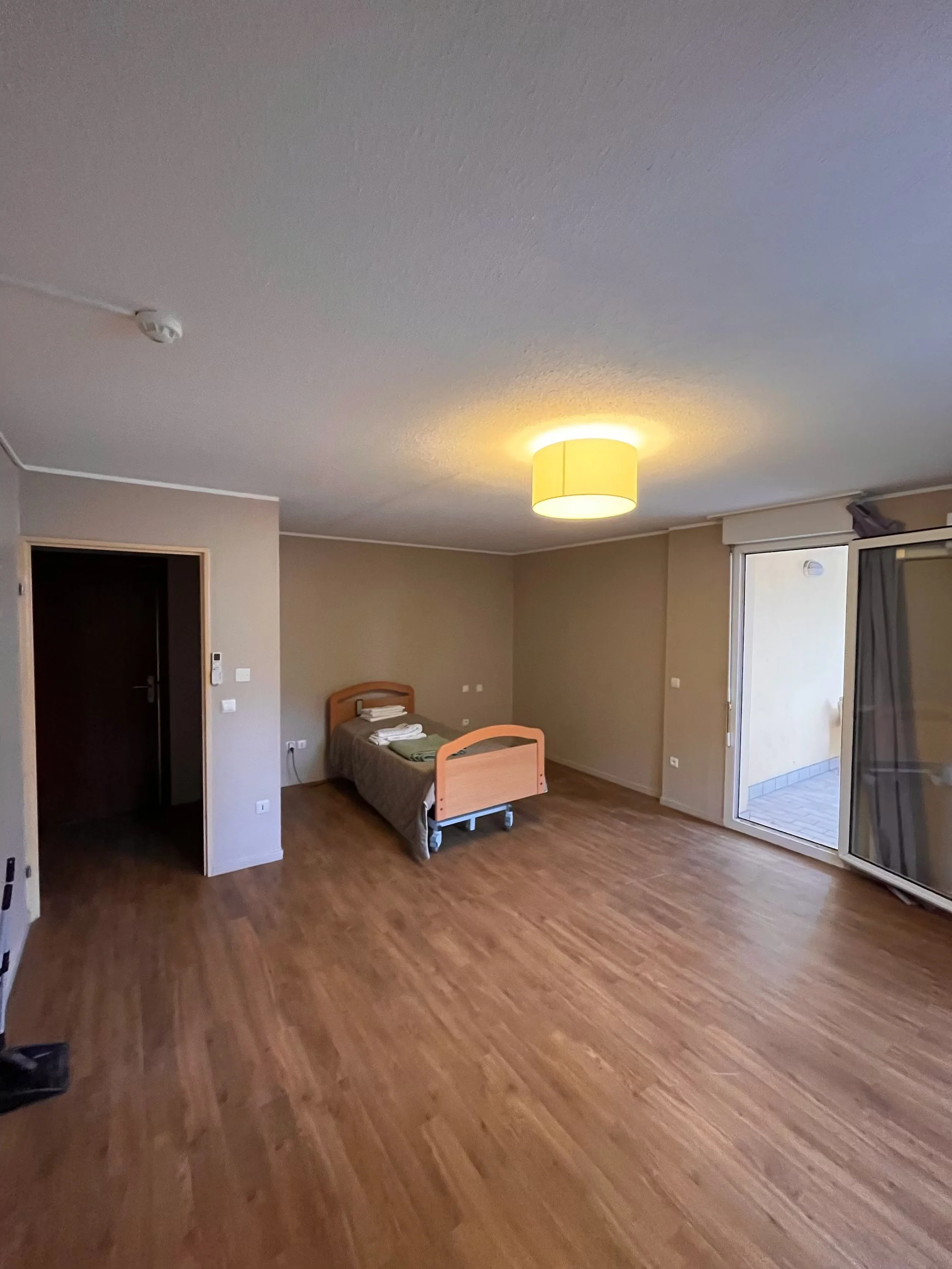 Chambre d'hôtel vide avec un lit simple, un balcon, et un sol en bois.