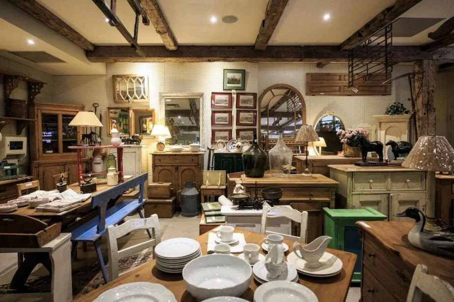 Une pièce meublée avec des articles de décoration vintage, comprenant des tables en bois, des lampes, des vases, des cadres photo et de la vaisselle, semblant être une boutique ou une aire d'exposition d'antiquités.