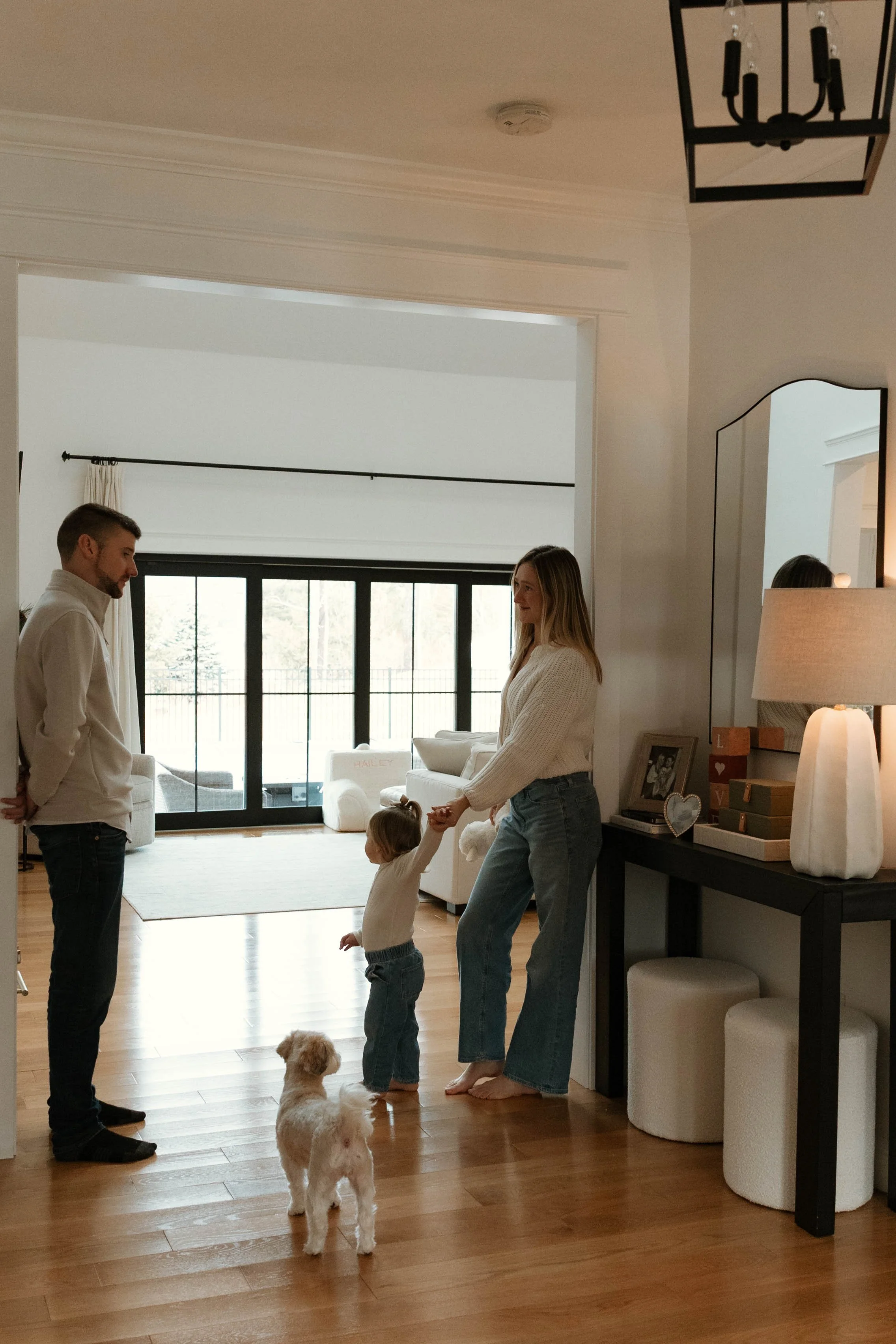 A family of four and a dog inside a living room. A woman is holding a child's hand, both are standing and smiling, facing a man who is standing with hands in pockets. The living room has large glass doors and natural light, with modern furniture and household decor visible.