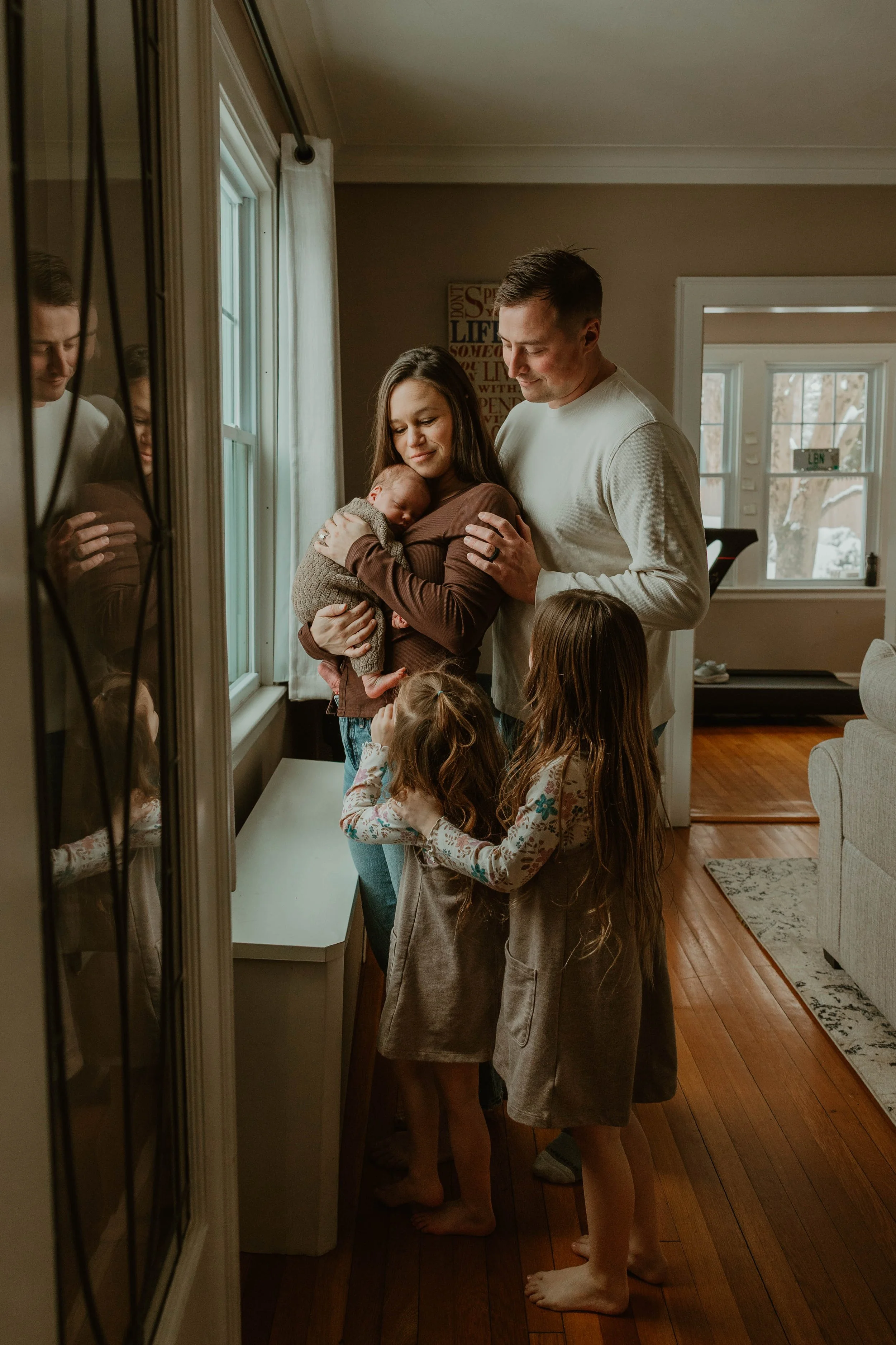 Family gathered near a window, with a woman holding a newborn baby, and two young girls hugging her. The man stands close, smiling. The scene is warm and intimate in a cozy home.