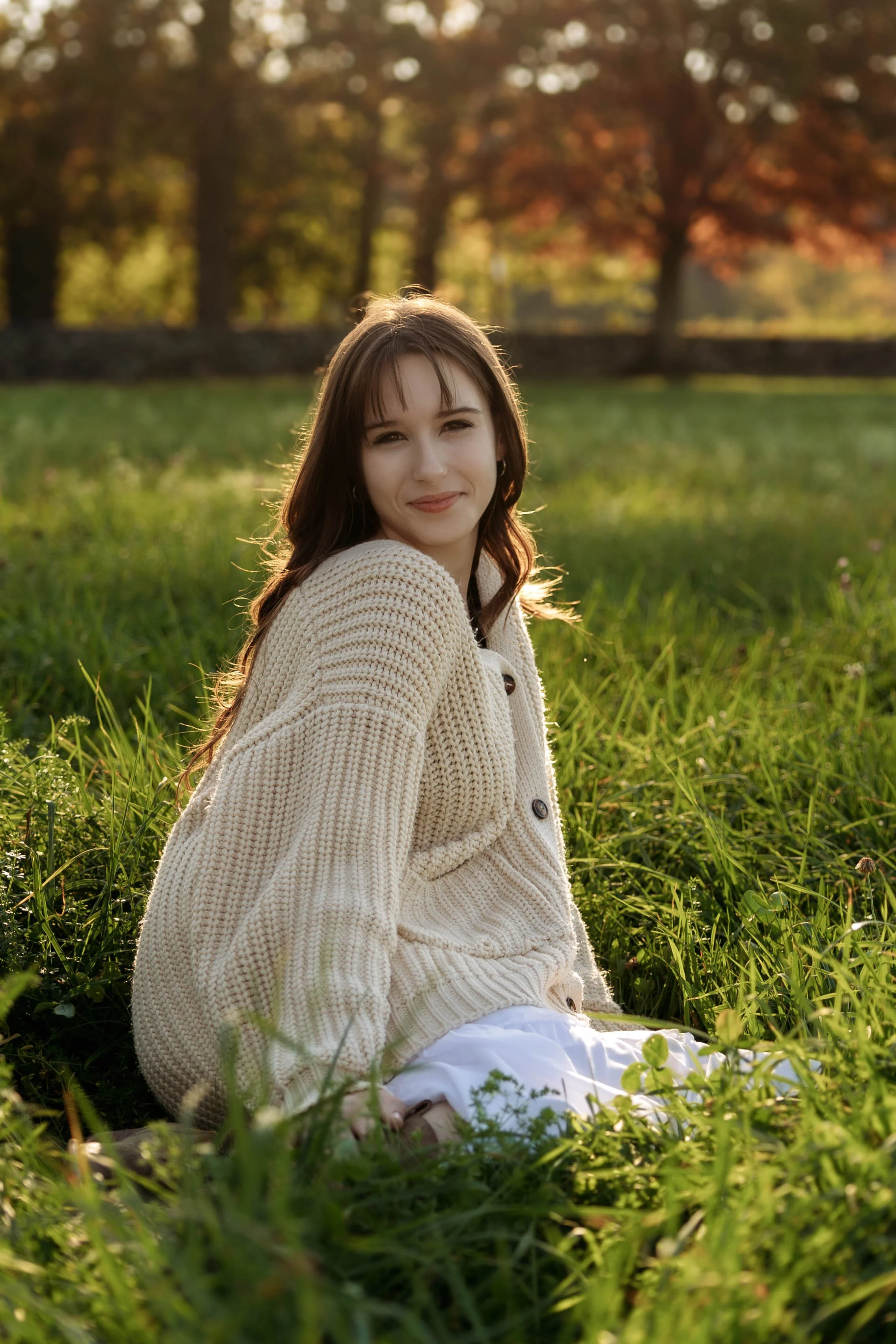 A young woman sitting in a grassy field during sunset, wearing a cream-colored cardigan and white dress, smiling at the camera.