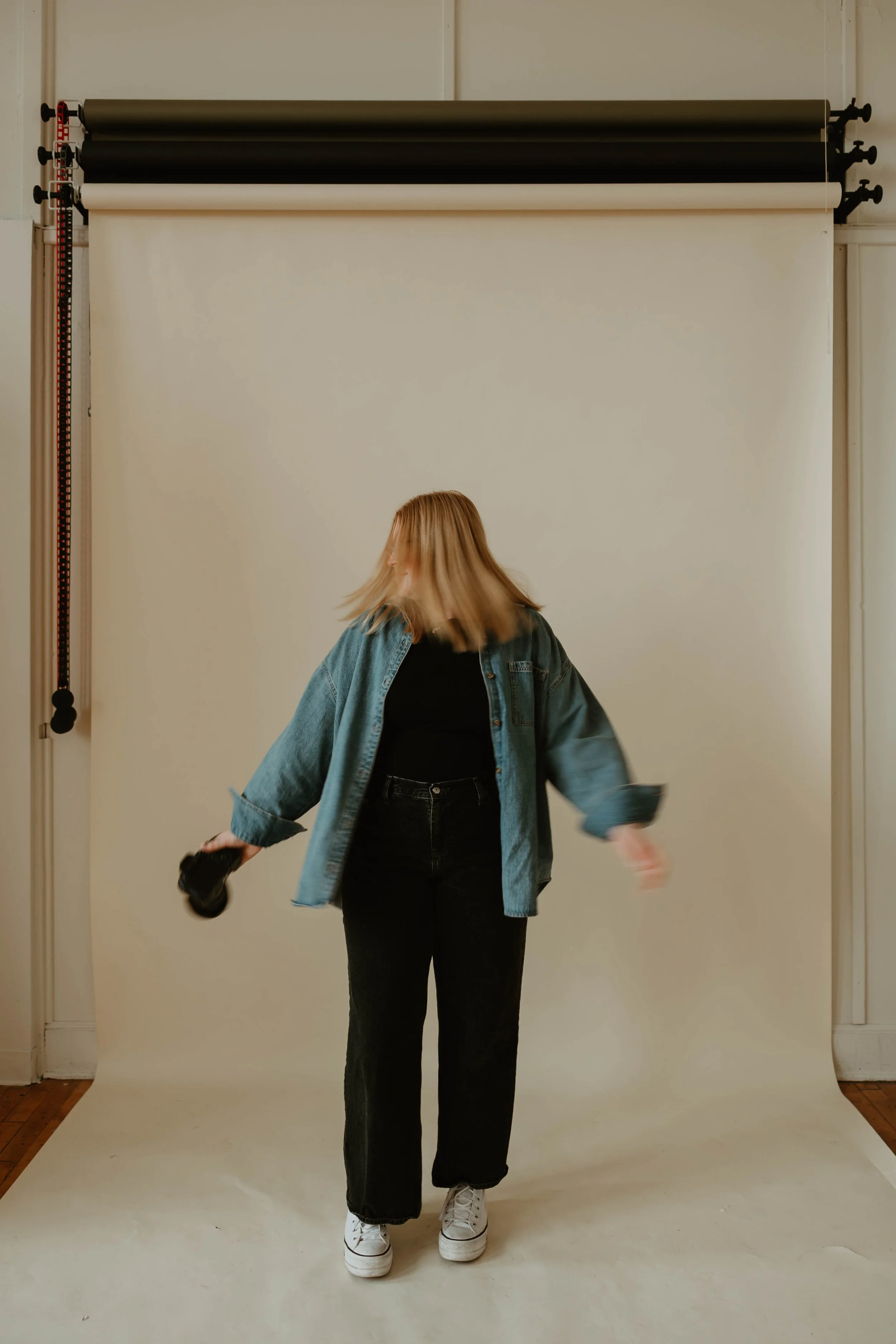 A person with blonde hair wearing a denim jacket, black shirt, black pants, and white sneakers, holding a camera, standing in front of a photography backdrop.