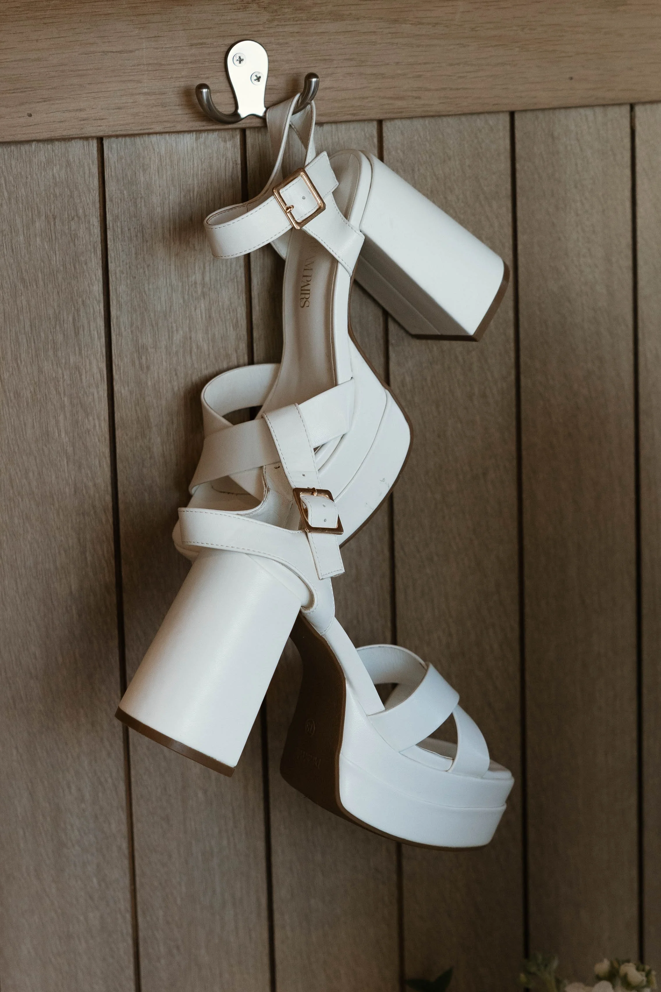 White high-heeled sandals hanging on a hook against a wooden wall.