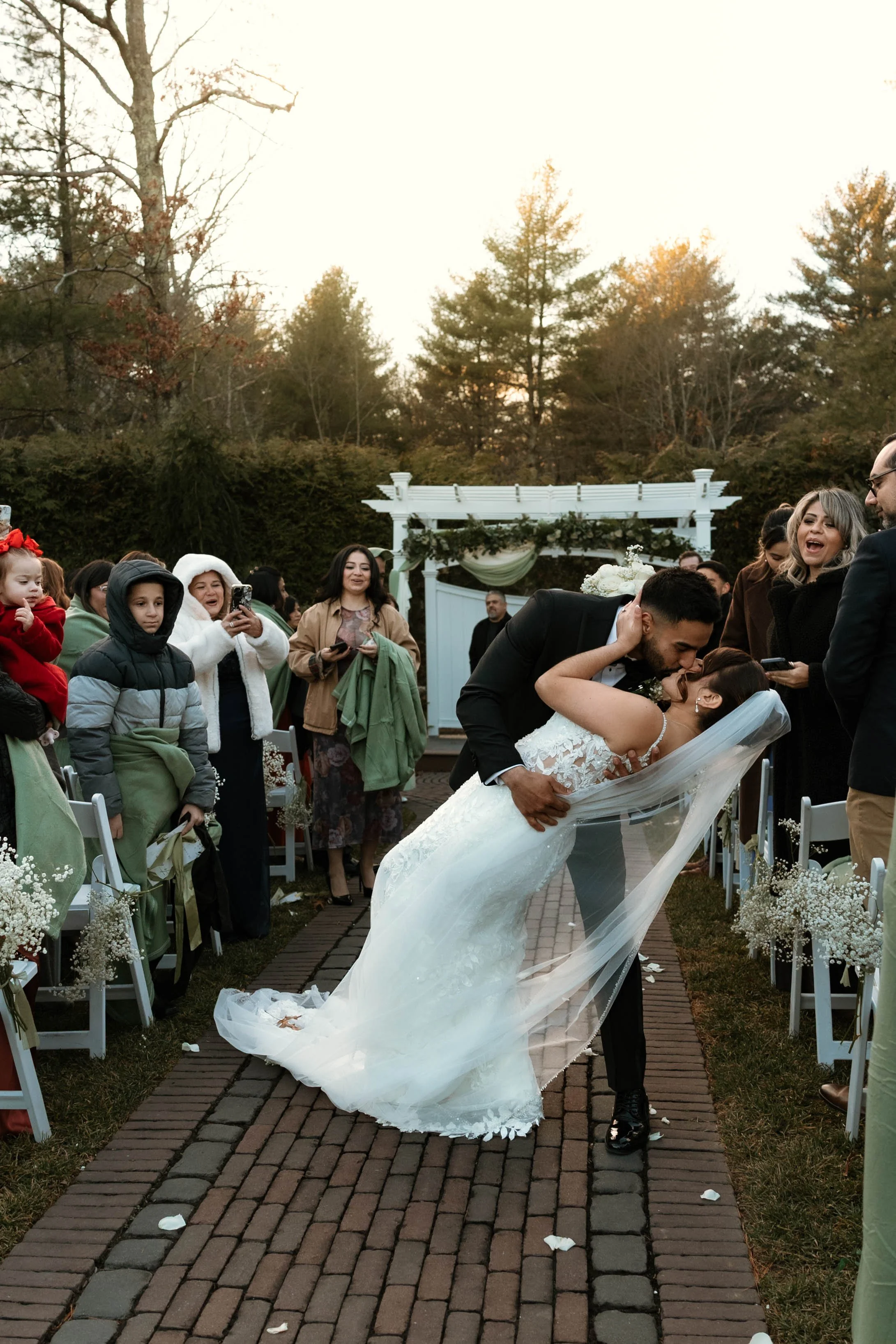A newlywed couple shares a kiss during their outdoor wedding ceremony, surrounded by friends and family, in the late afternoon with trees in the background.