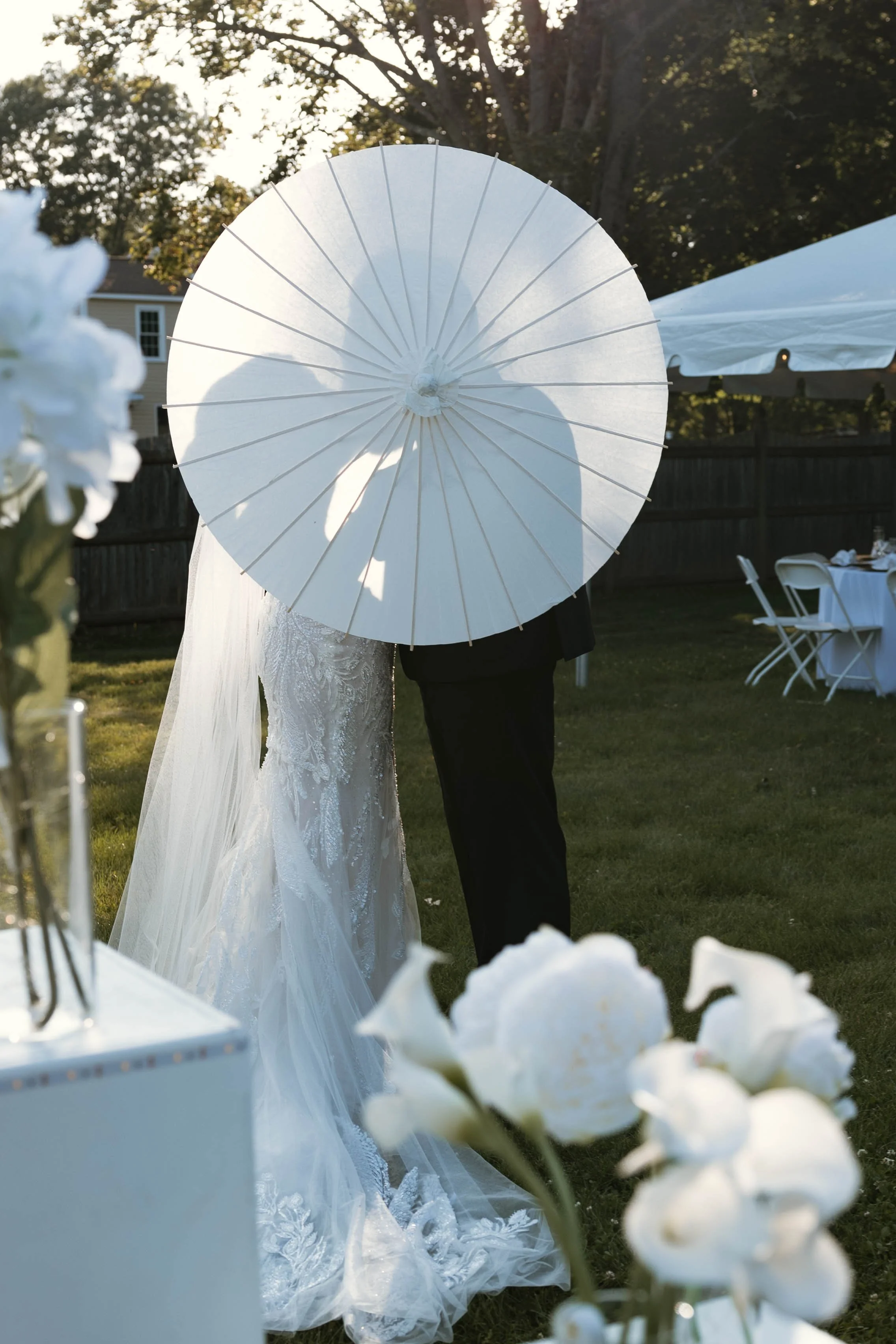 A bride and groom standing outdoors at a wedding reception, with the bride in a lace gown and the groom in a black suit, holding a white parasol that covers their faces. Flowers and tables are visible in the background.