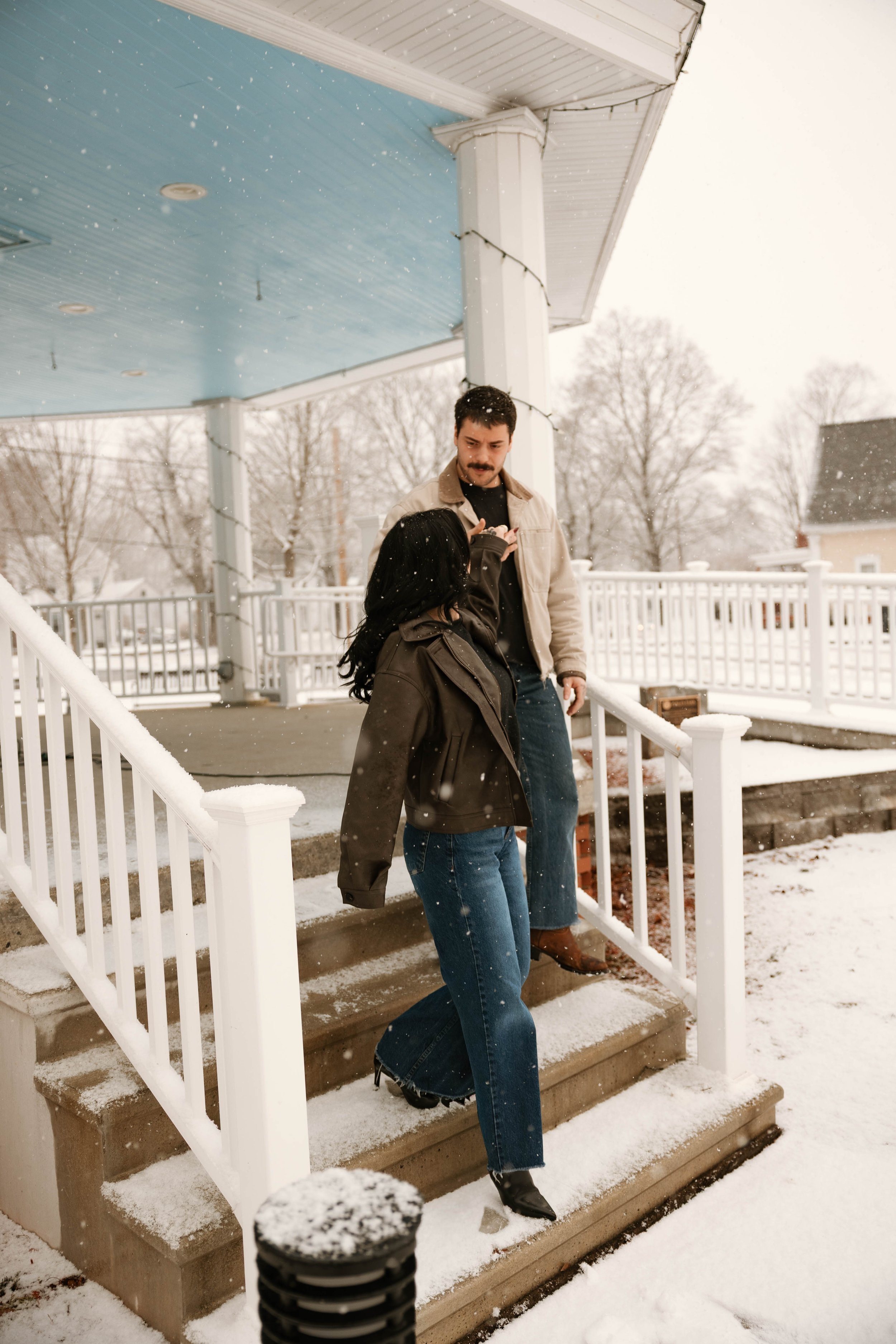 A man and woman standing on snowy porch steps outside a house during snowfall. The man is looking at his phone, the woman is looking toward the steps. Both are dressed in winter clothing.