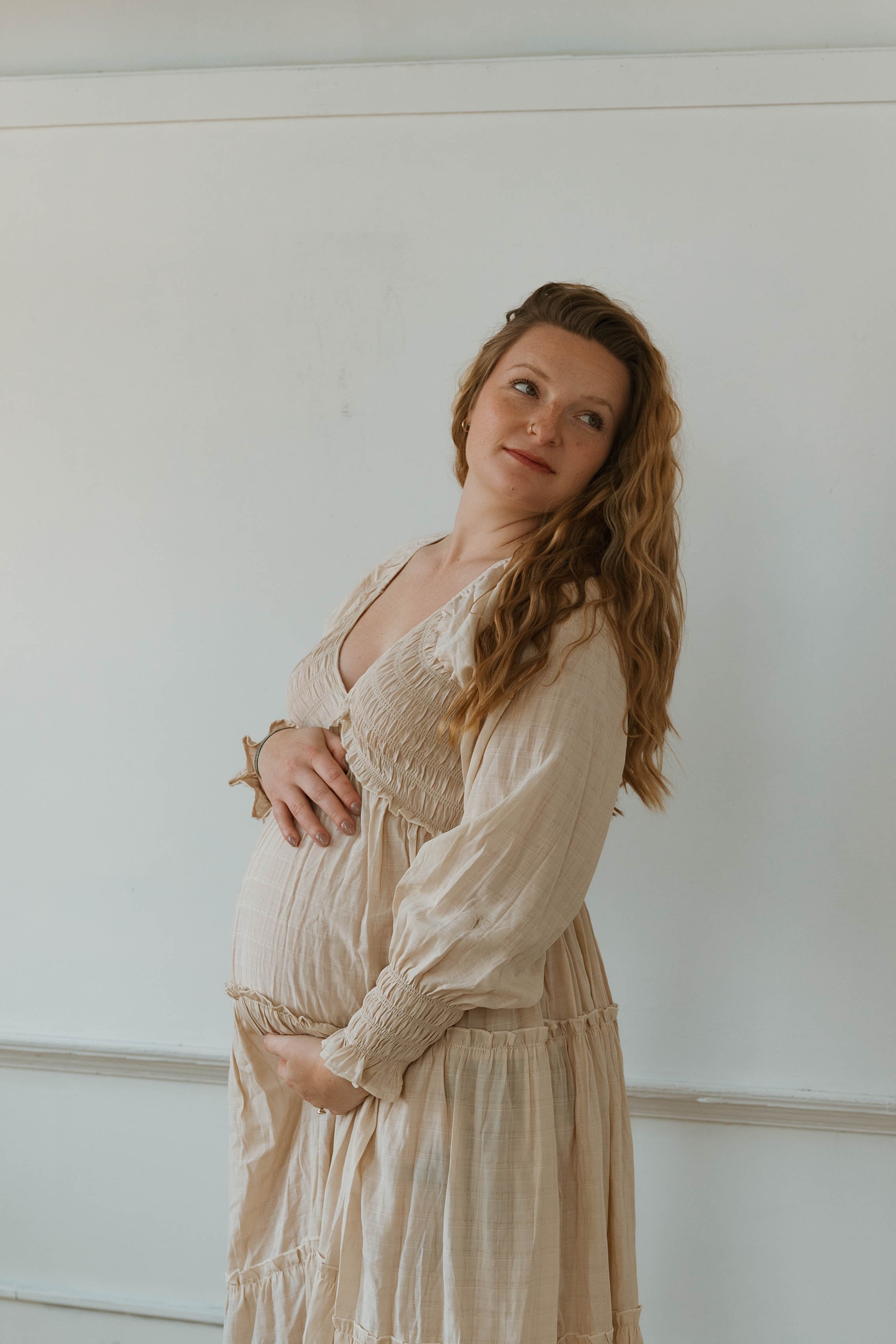 Pregnant woman with long red hair in a beige dress standing against a white wall.