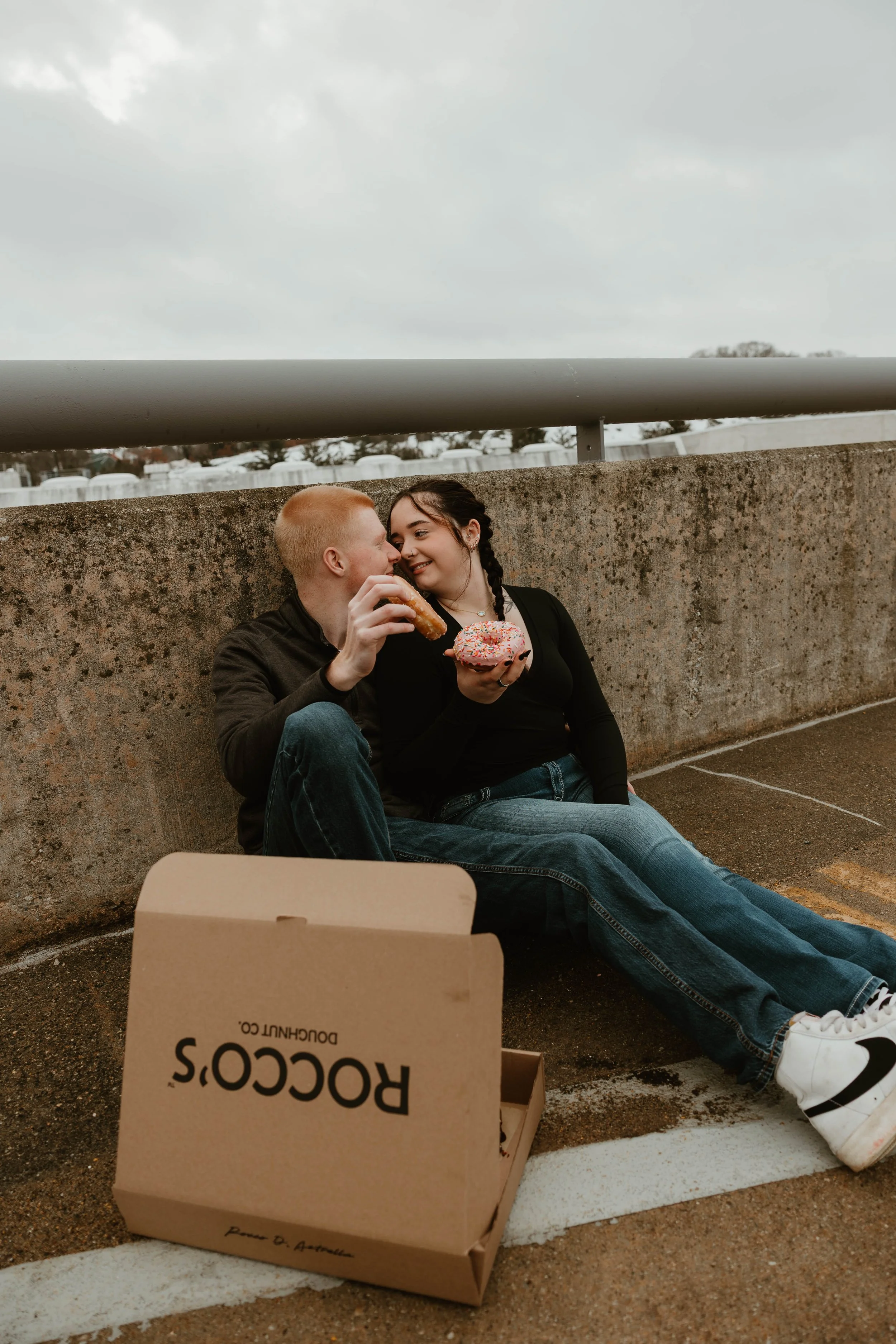 A young couple sitting on the ground beside a concrete wall, sharing a moment with pizza and donuts. There is an open pizza box in front of them with the Rocco's Doughnut Co. logo visible.