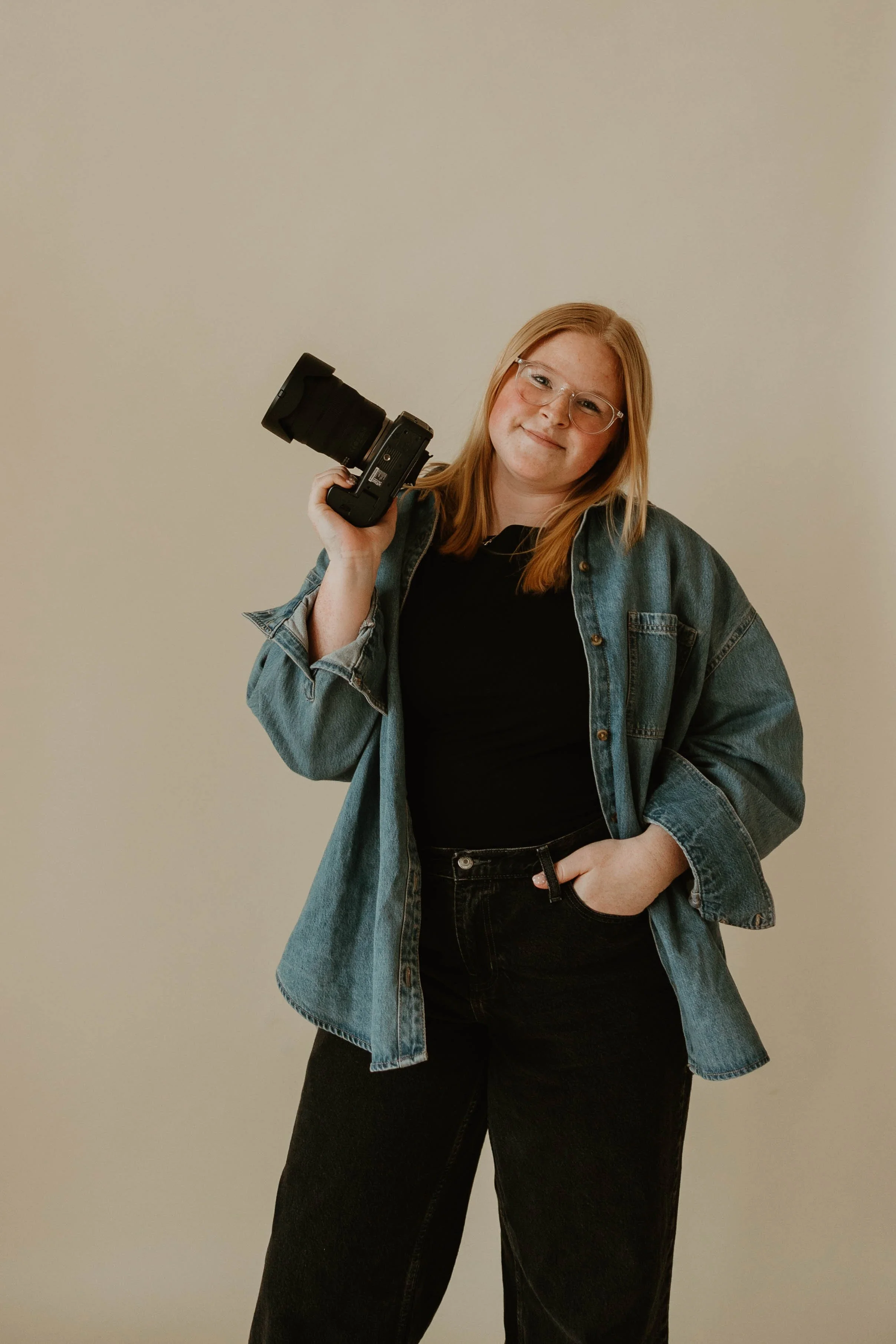 A young woman with red hair and glasses smiling while holding a camera over her shoulder, wearing a denim jacket and black pants, standing against a plain beige wall.