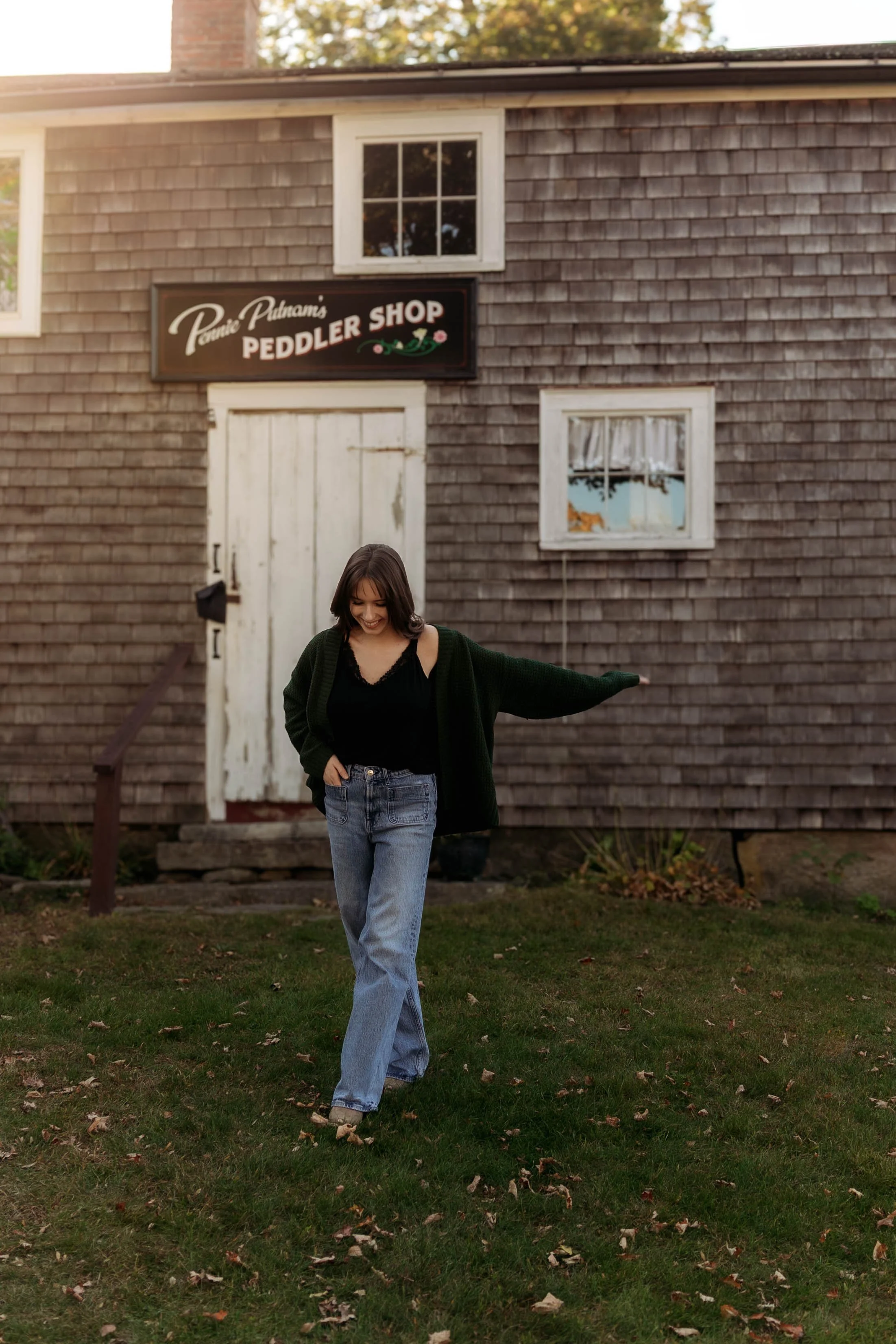 A woman standing in front of a rustic wooden building with a sign that reads 'Penu Pine's Peddler Shop', smiling and looking down, with one arm extended outward and one hand in her pocket, wearing jeans, a black top, and a dark green cardigan.