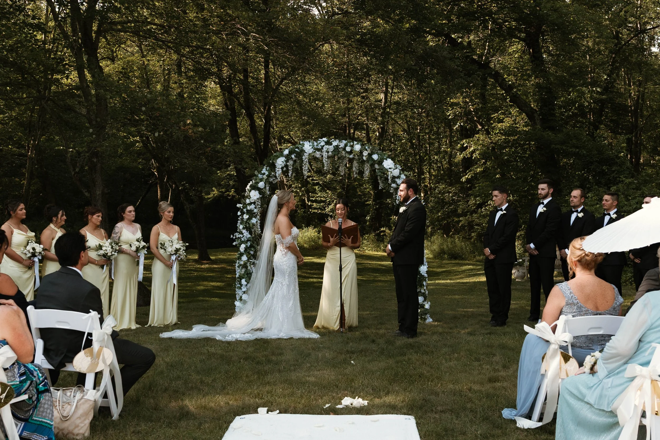 A wedding ceremony taking place outdoors in a wooded area, with the bride and groom standing under a flower arch, surrounded by bridesmaids and groomsmen, guests seated nearby, and a officiant reading from a book.
