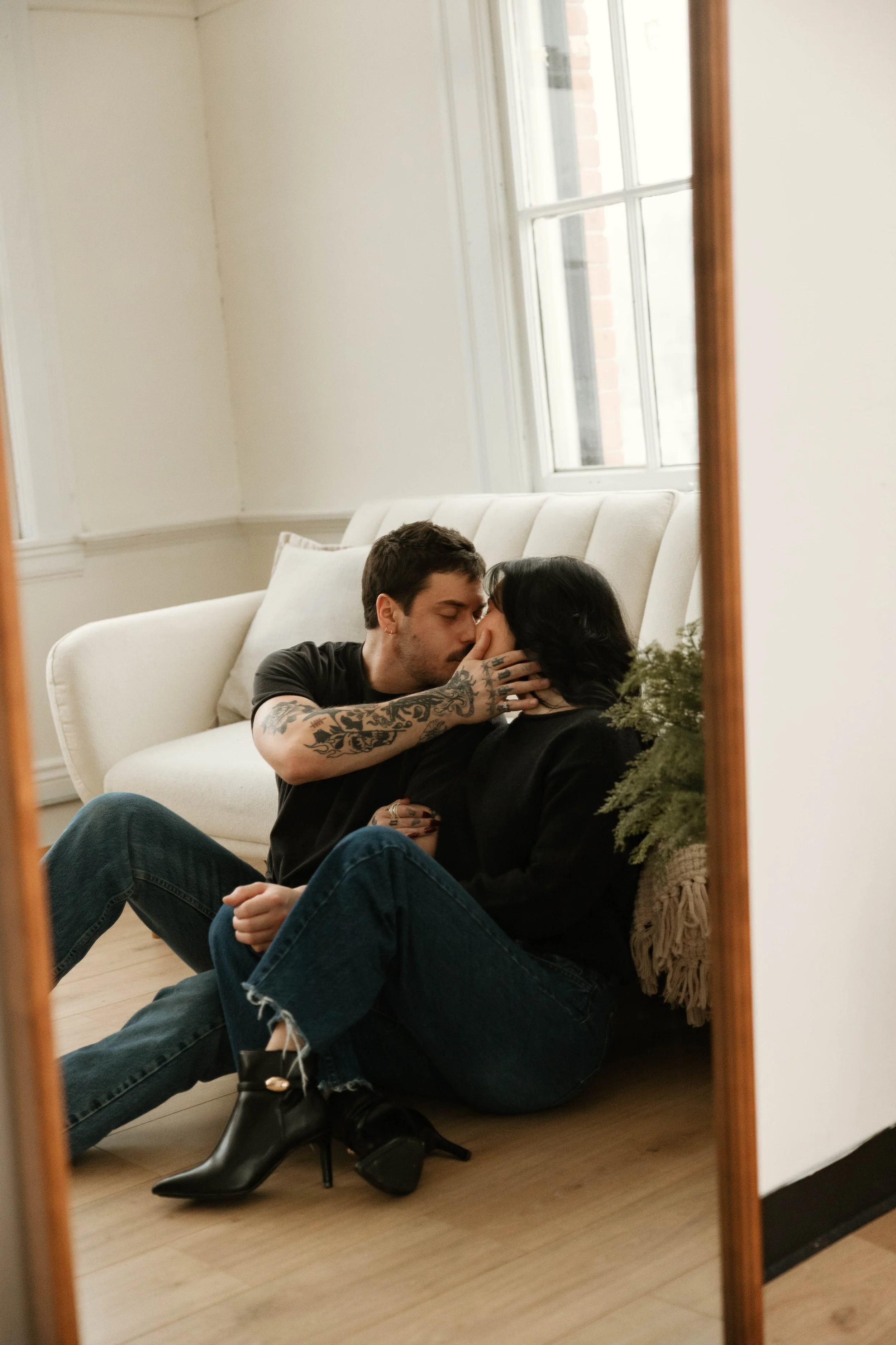 A couple sharing an intimate moment on the floor in front of a mirror, with a white couch and window behind them.