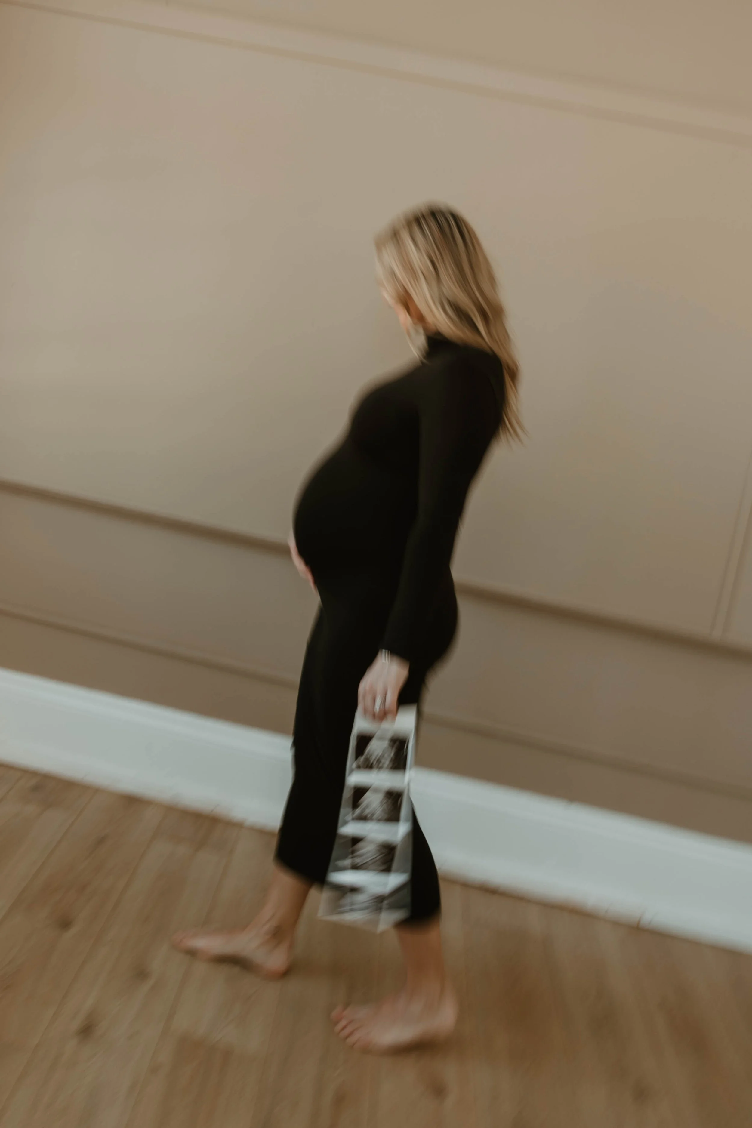 Pregnant woman with blonde hair wearing black clothing, holding an ultrasound photo, walking barefoot on a wooden floor in a room with beige walls.