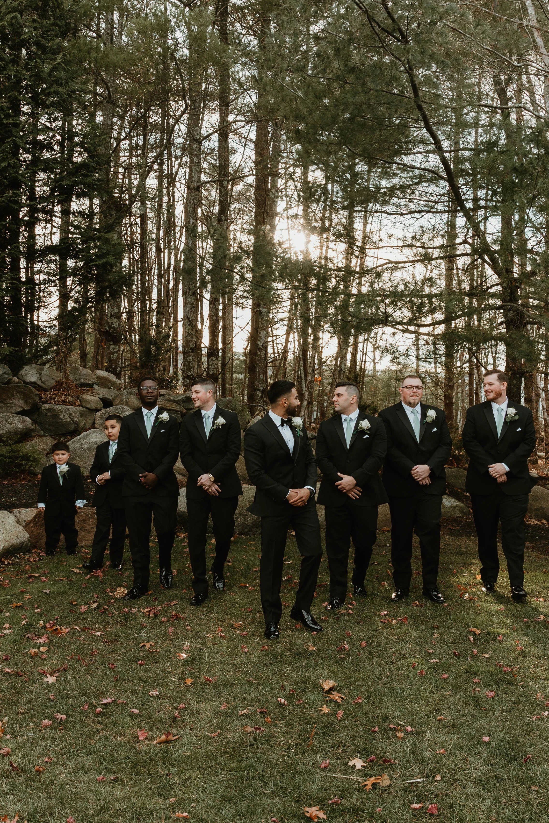 A group of seven groomsmen and two boys dressed in tuxedos walking outdoors on a grassy area with trees in the background during sunset.