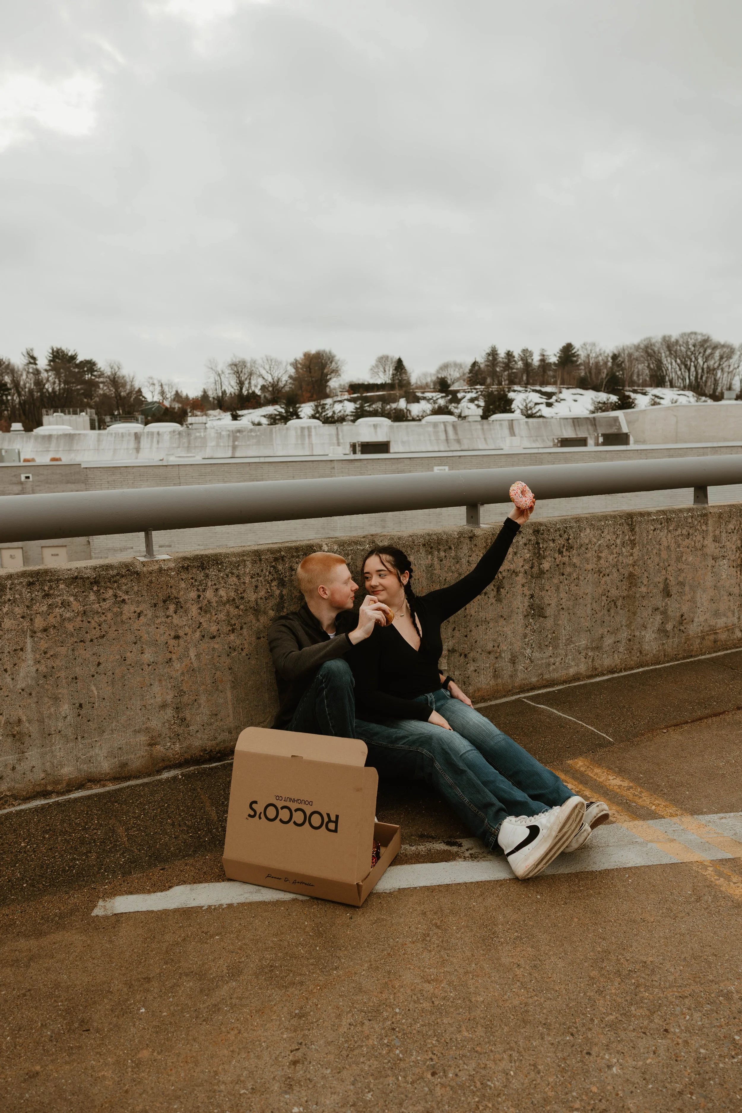 A young couple sitting on the ground near a parking lot railing, with a pizza box and a donut, sharing a moment.