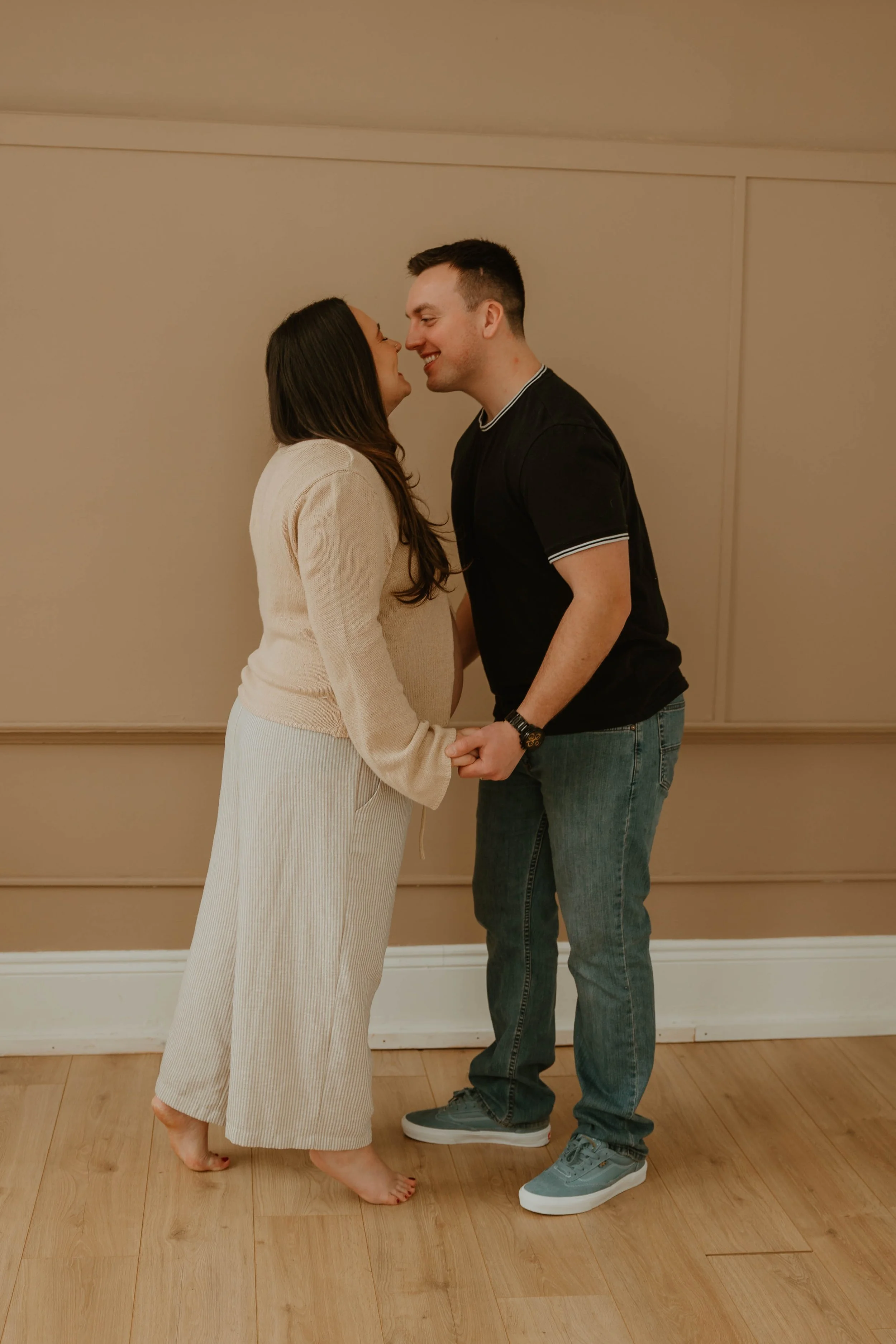 A couple holding hands and smiling at each other, standing close together indoors on a wooden floor with beige walls.