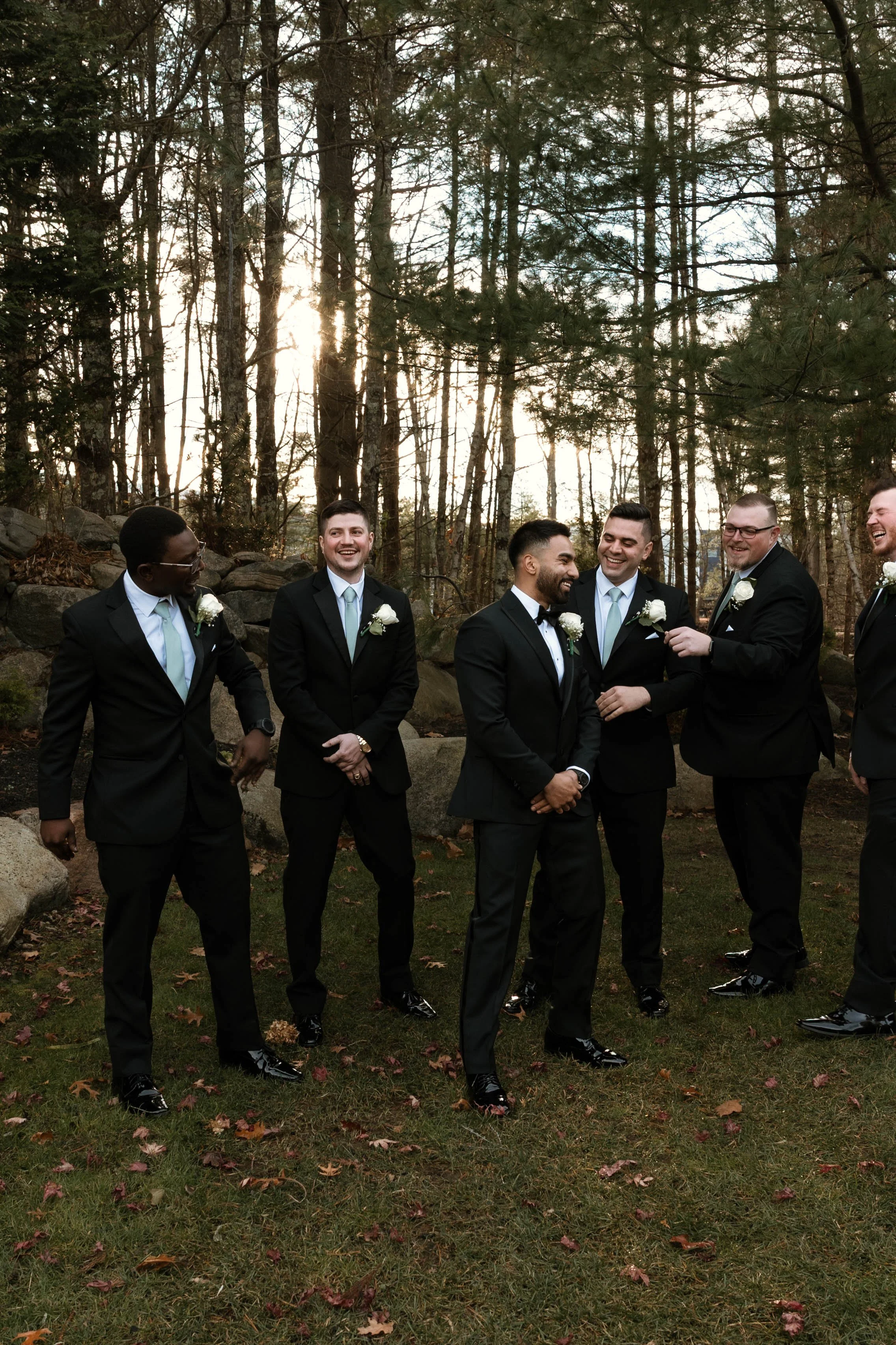 A group of men in black suits with white shirts and light blue ties, standing outdoors on grass with fallen leaves, surrounded by trees during sunset, all smiling and laughing.