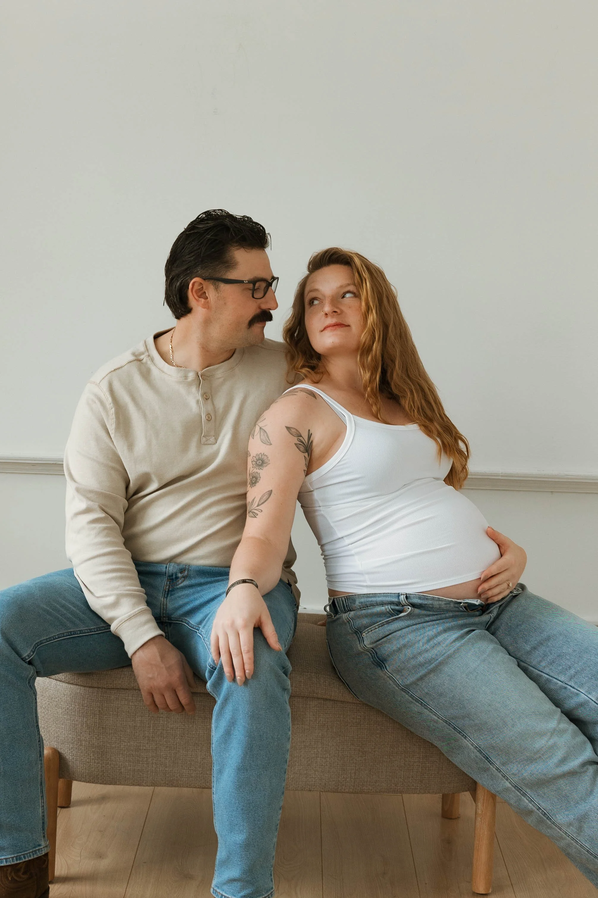 A pregnant woman with long, wavy red hair, wearing a white tank top and jeans, sitting on a bench, touching her belly, looking at a man with glasses and a mustache, wearing a beige shirt and jeans, sitting beside her, who is looking at her