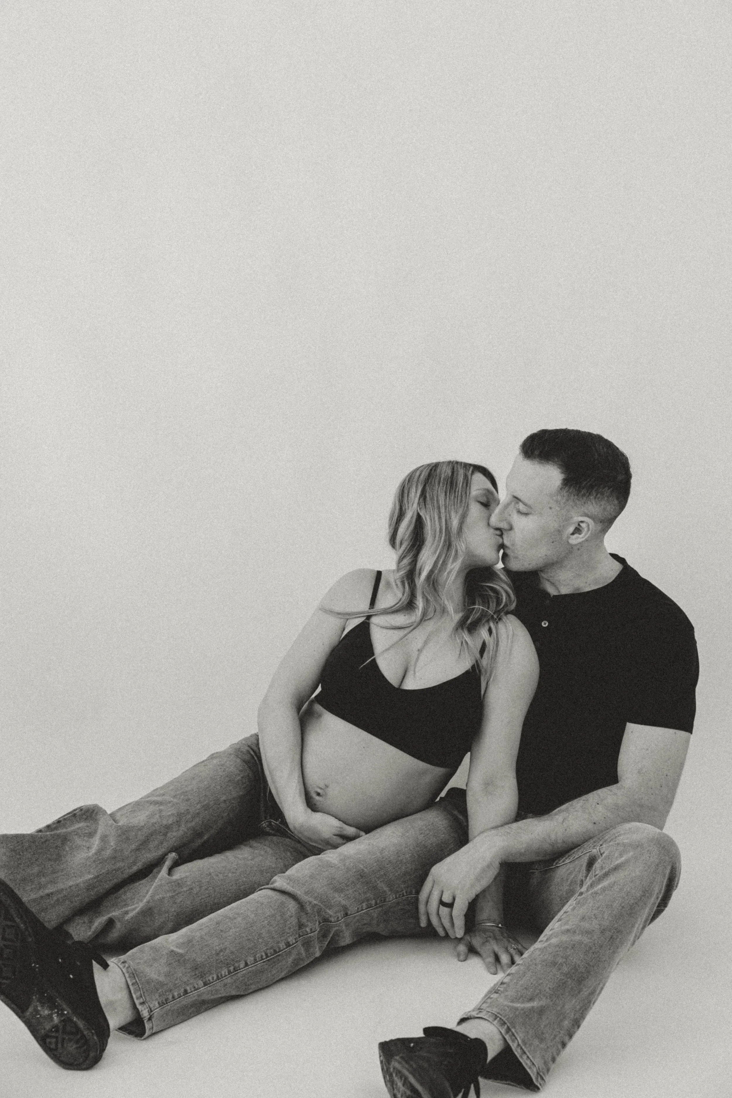 A pregnant woman and a man sit on the floor, kissing, with the woman gently resting her hand on her belly.