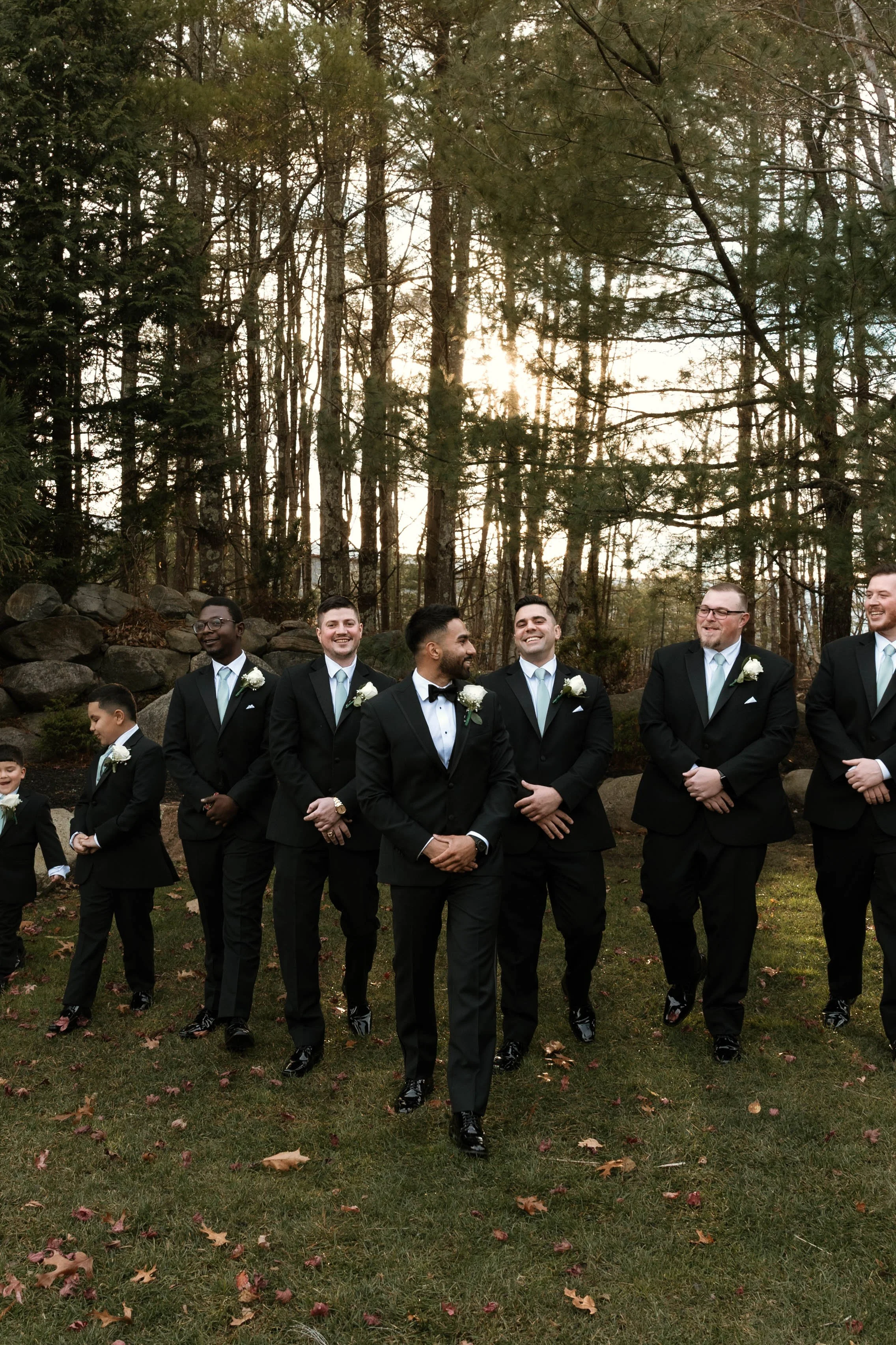 A group of men and boys in formal suits walking outdoors in a wooded area, smiling and laughing together.
