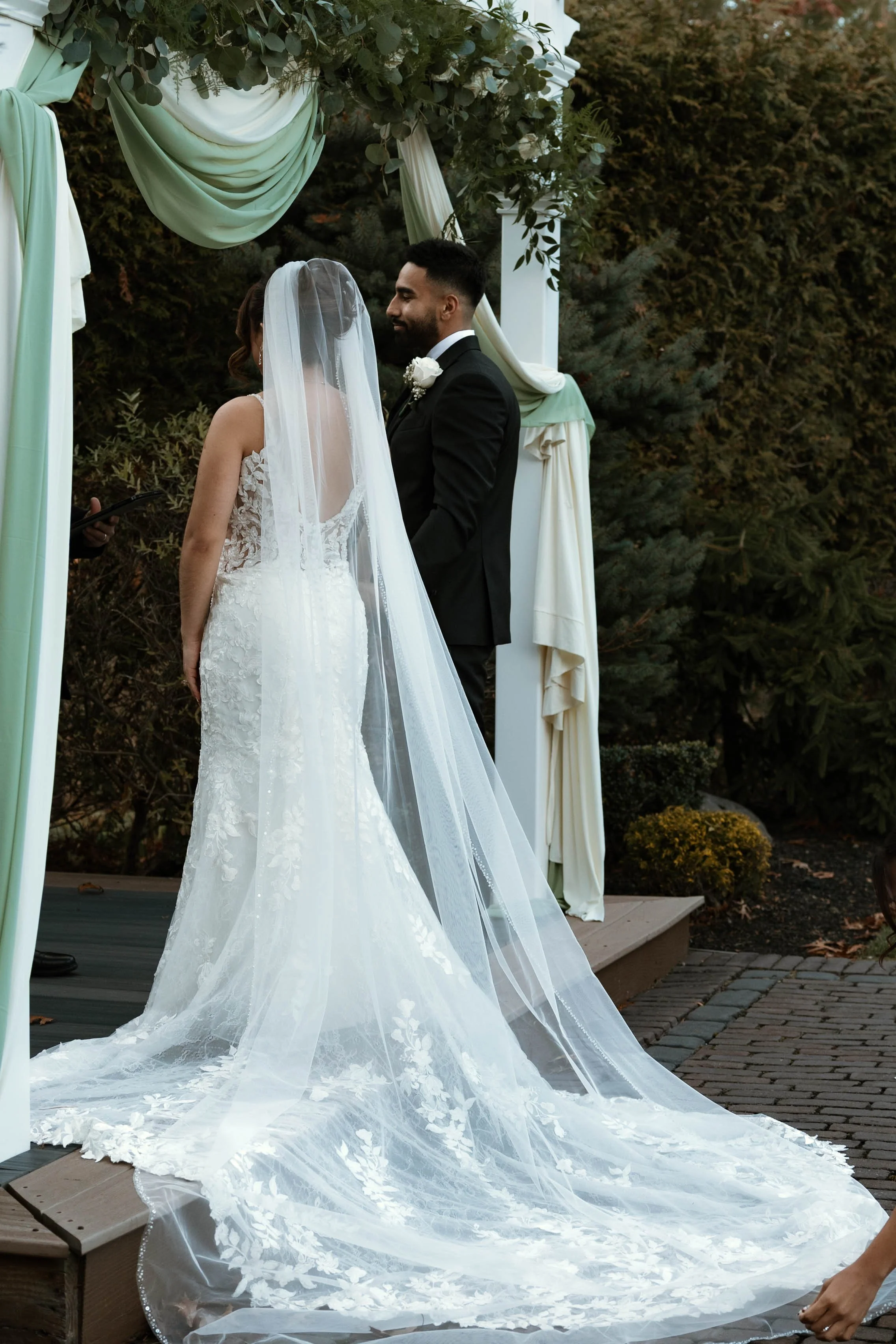 A bride and groom exchanging vows during an outdoor wedding ceremony, with the bride wearing a lace wedding gown and veil, and the groom in a black suit, standing under a decorated archway surrounded by greenery.
