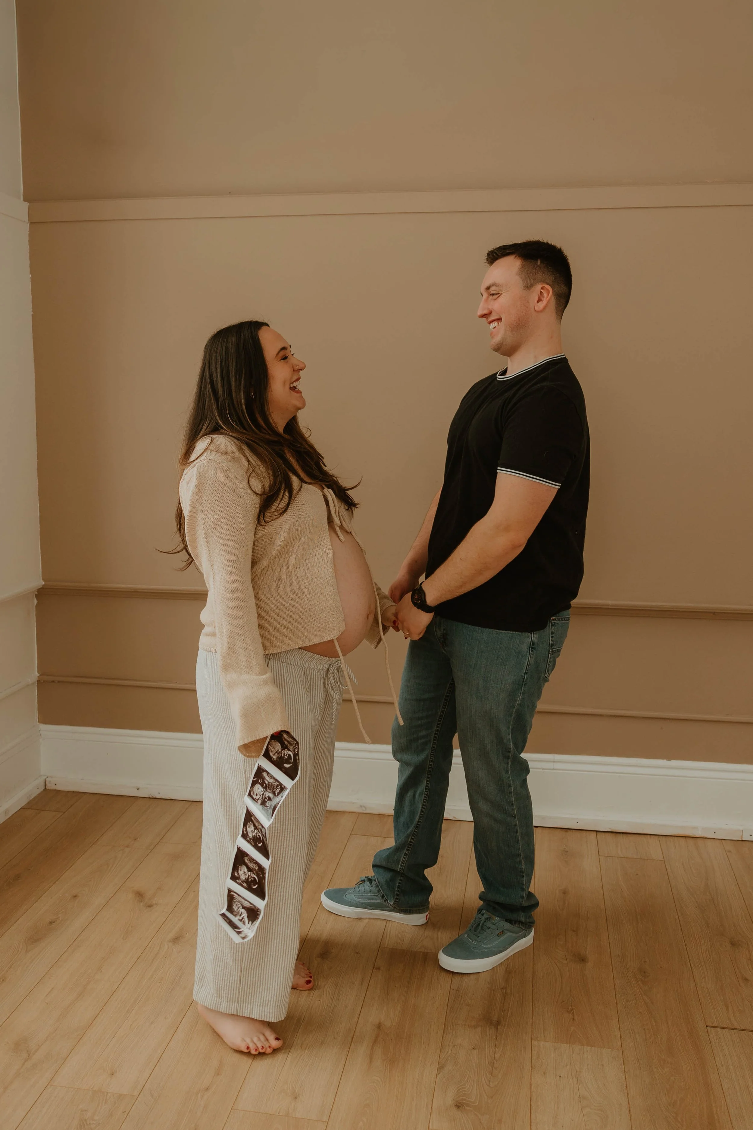 A smiling pregnant woman holding ultrasound pictures standing in front of a smiling man, both holding hands indoors with light brown walls and wooden flooring.