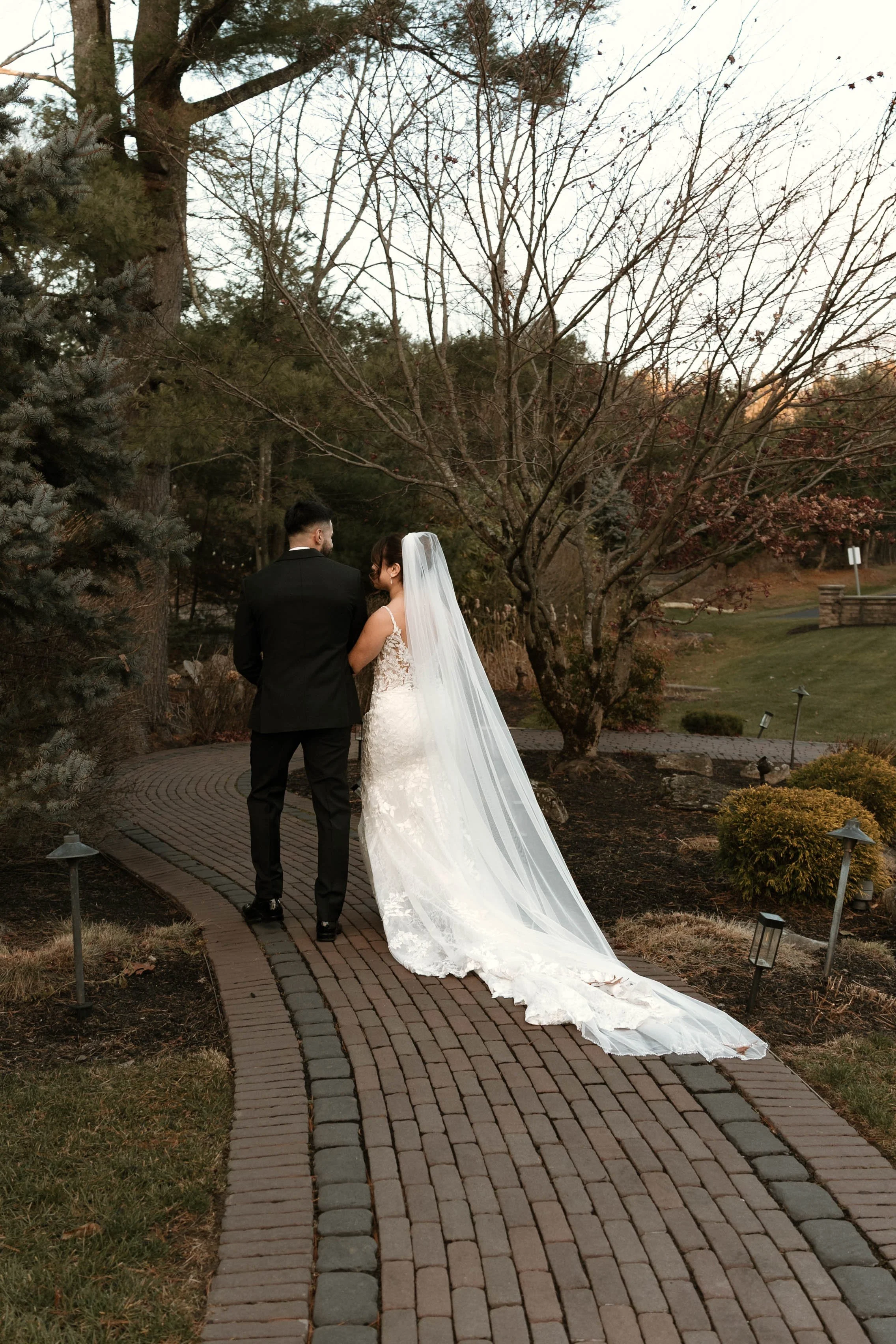 Bride and groom walking on a brick path in a park during sunset, with trees and shrubs around.