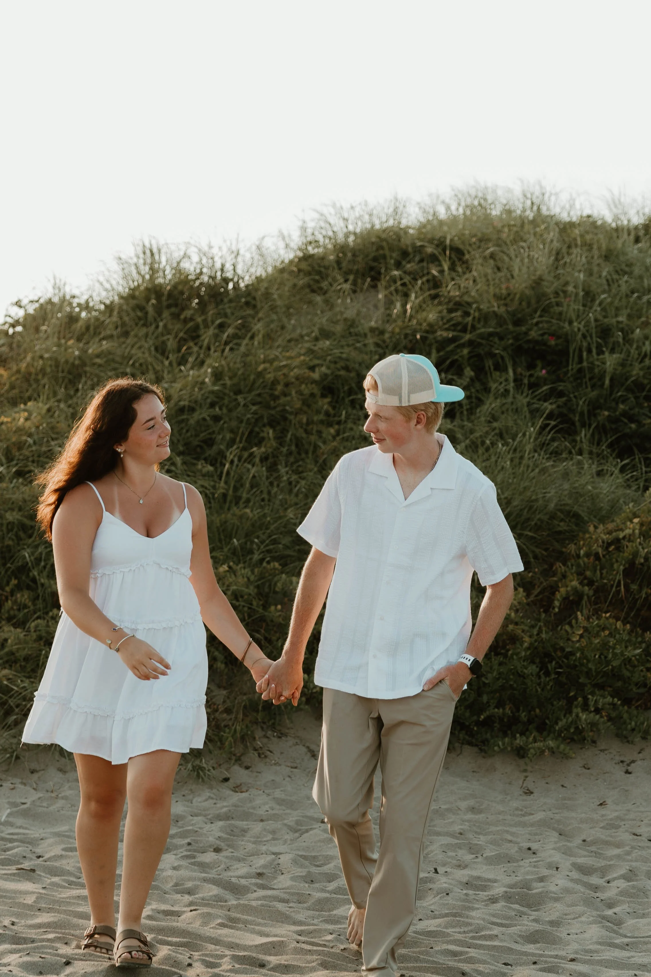 A young couple holding hands and walking on a sandy beach with grassy dunes in the background, enjoying a sunny day.