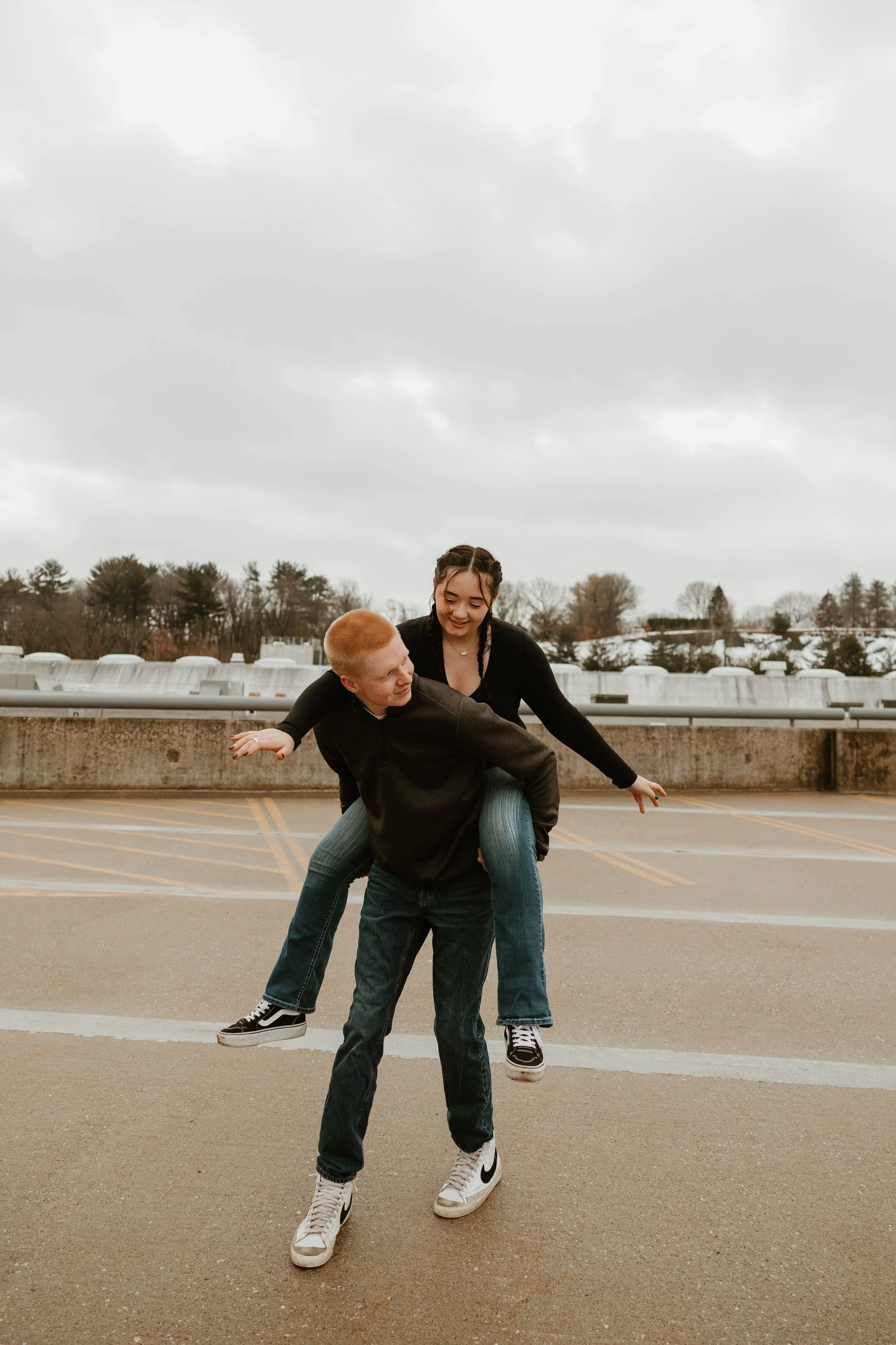 A boy giving a girl a piggyback ride in a parking lot on a cloudy day.