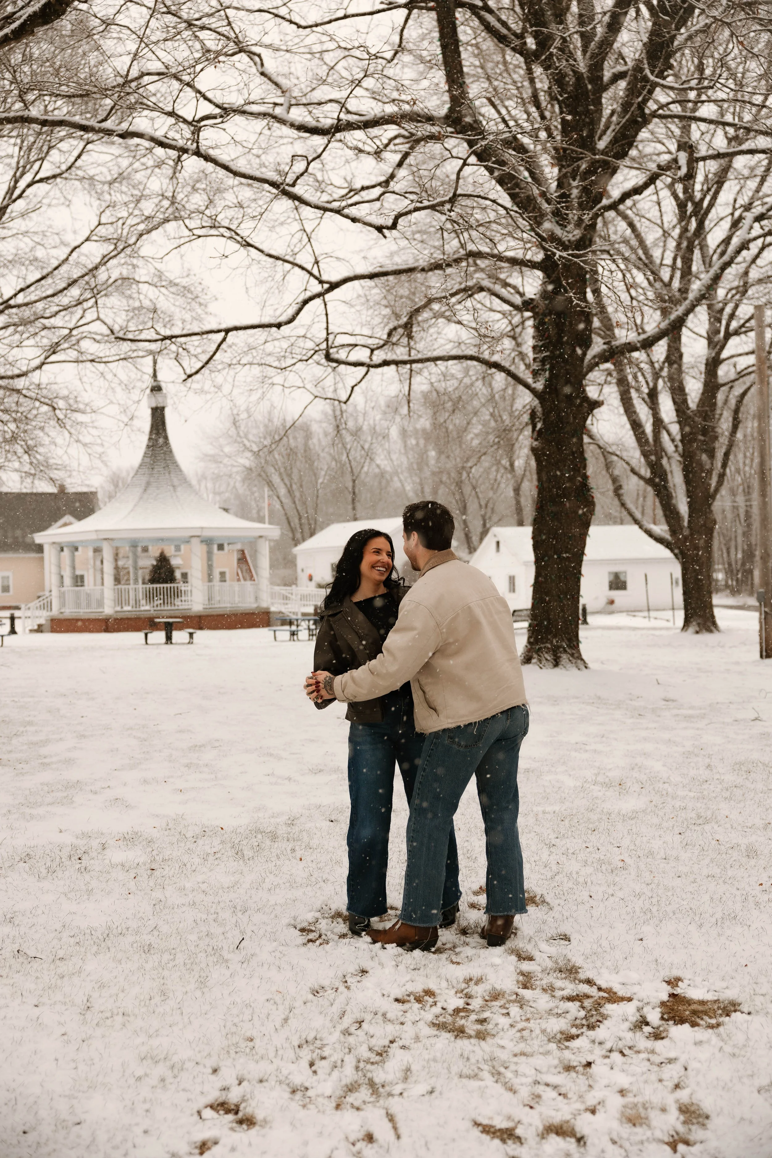 A couple dancing in the snow in a park with a gazebo and trees covered in snow.
