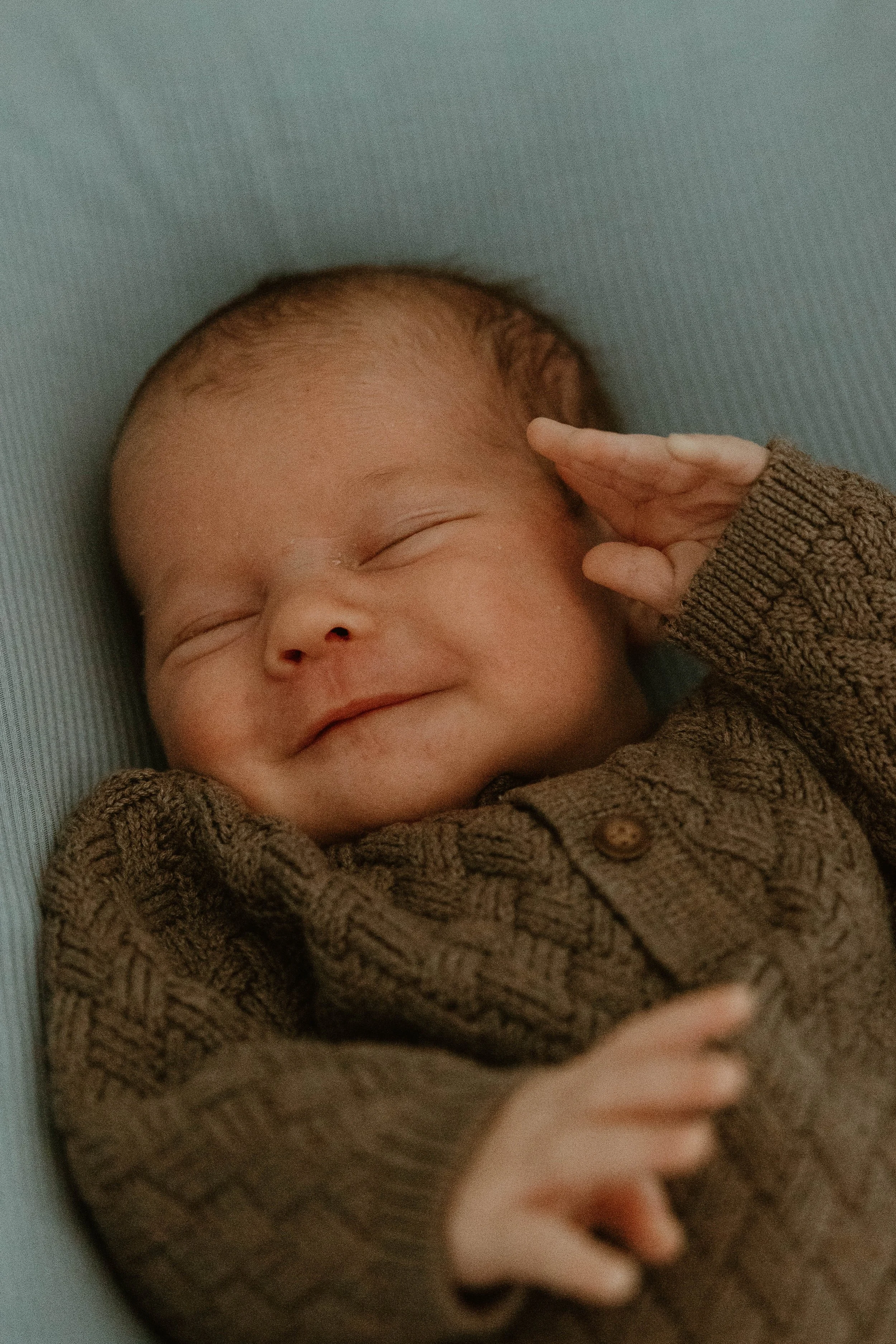 A smiling baby lying on a soft surface, wearing a brown knitted sweater, with one hand near their face and eyes closed.