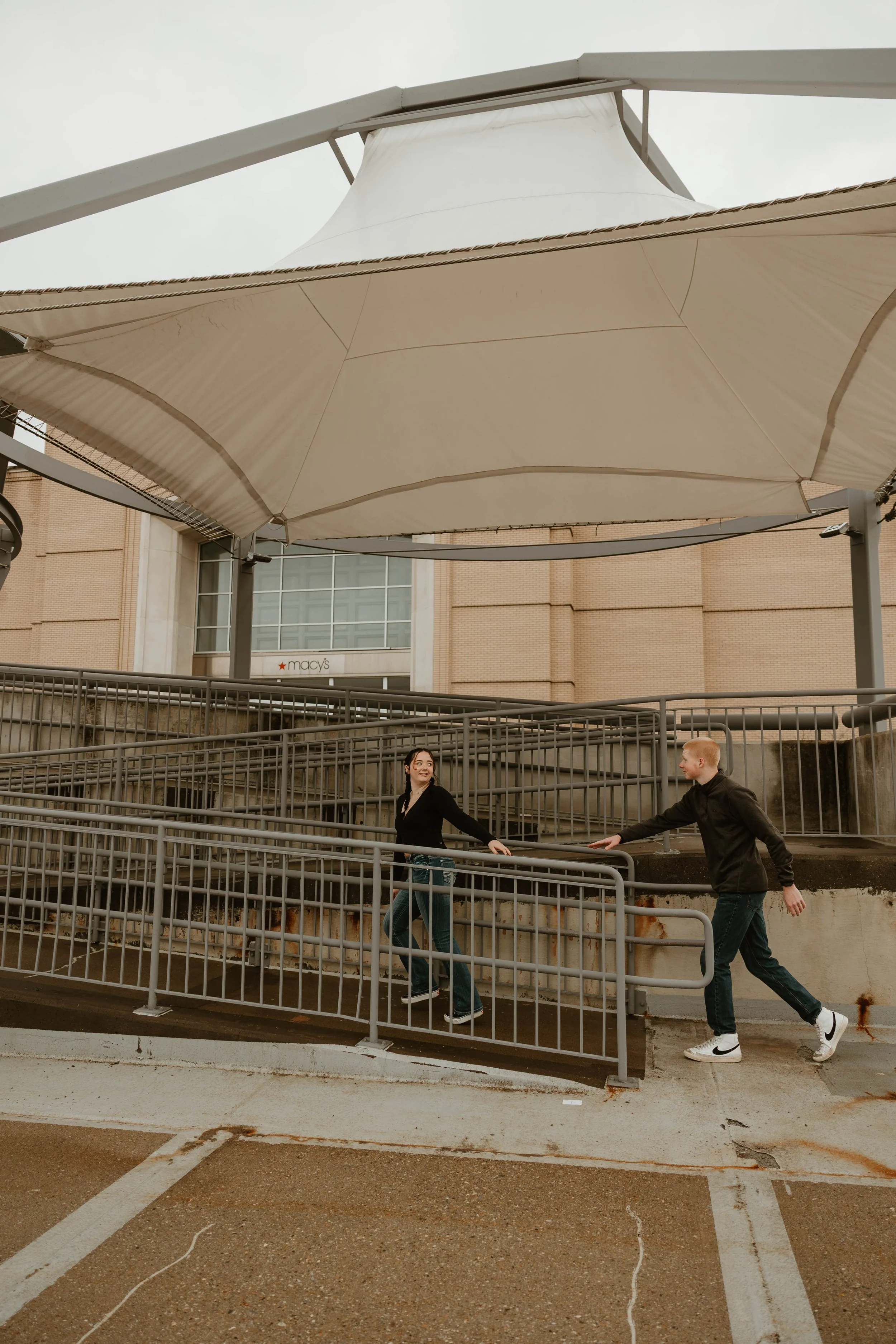Two teenagers, a girl and a boy, playing on a metal ramp at an outdoor shopping mall parking lot, with Macy's store visible in the background under a large fabric canopy.