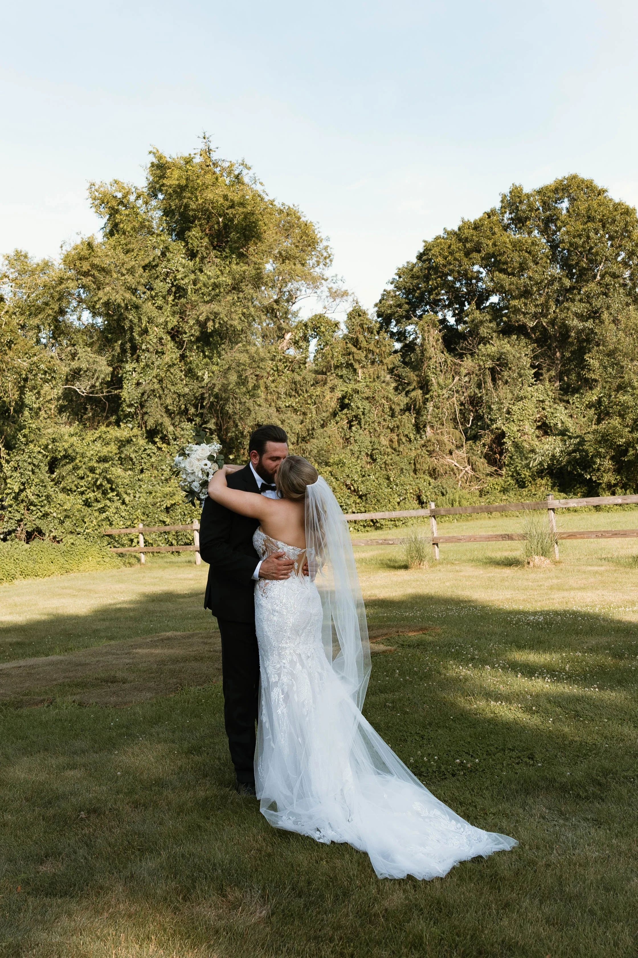 A bride and groom hugging outdoors on a grassy field with trees in the background, during daylight.