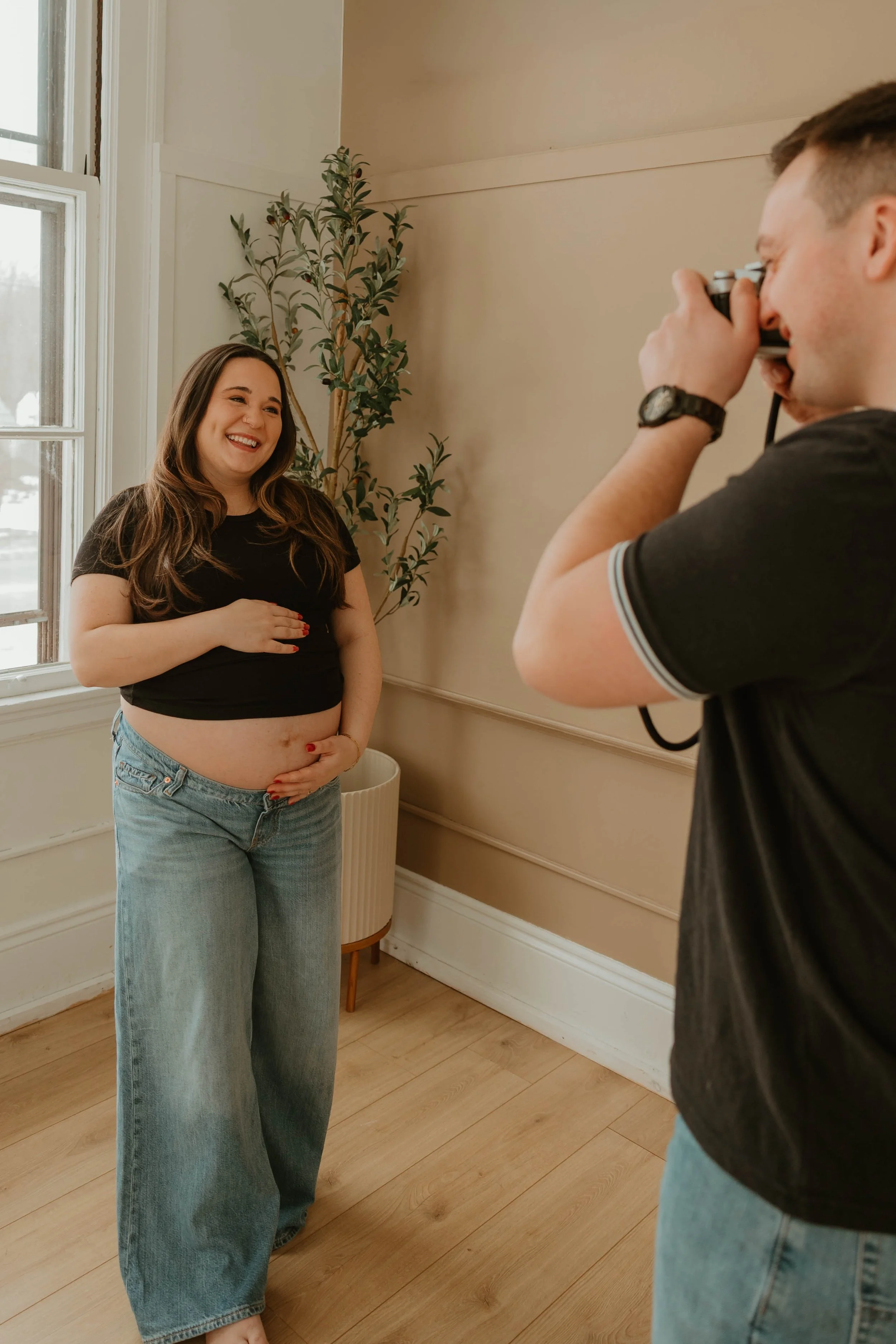 A woman is smiling and holding her belly as she gets ready for a photo shoot while a man with a camera takes her picture indoors near a window and a potted plant.