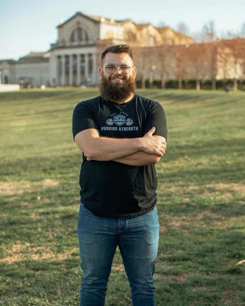 A man with glasses and a beard smiling and standing with his arms crossed on a grassy lawn with a large historic building in the background.