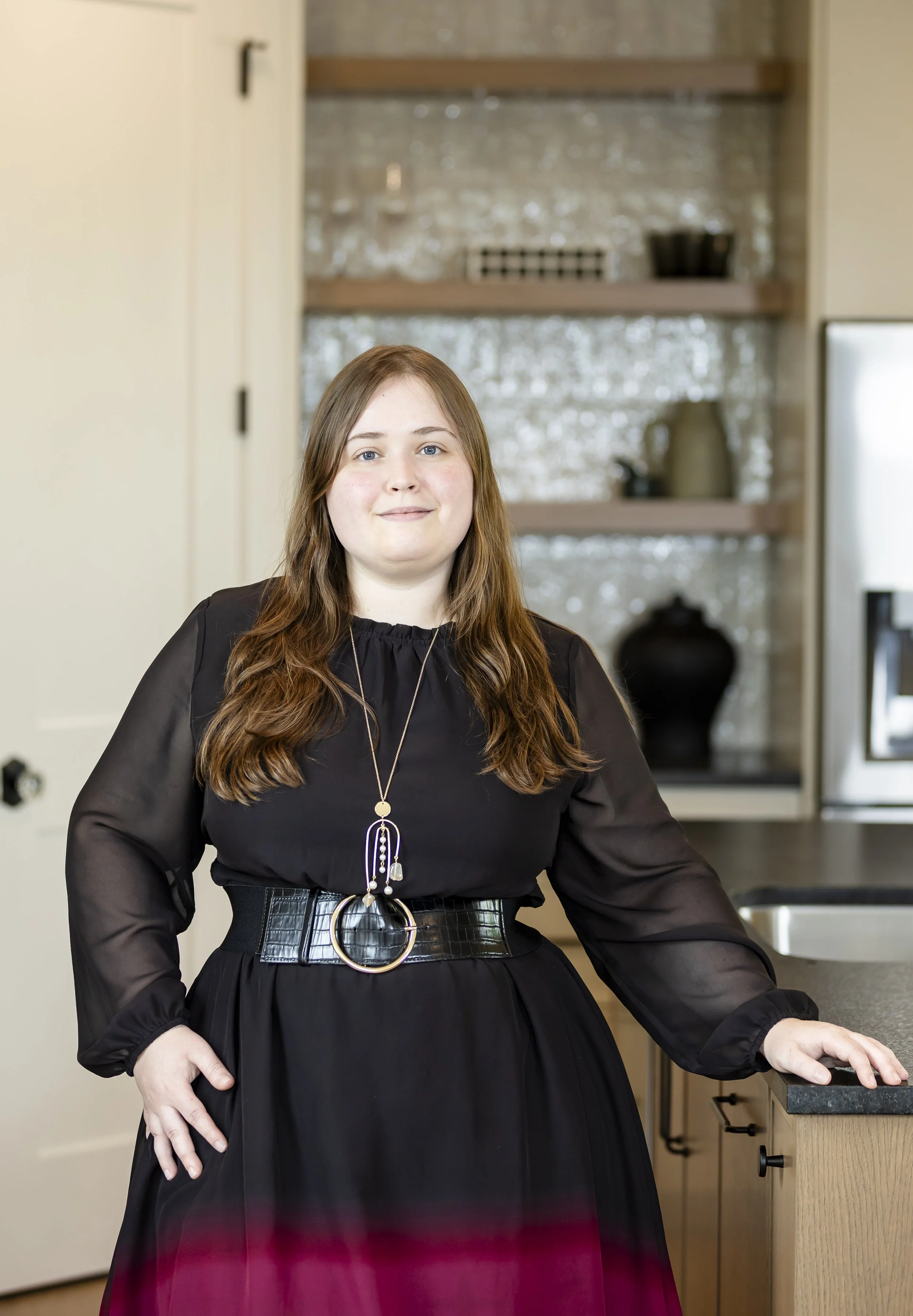 A woman with long brown hair, wearing a black dress with sheer sleeves and a wide black belt, standing in a modern kitchen.