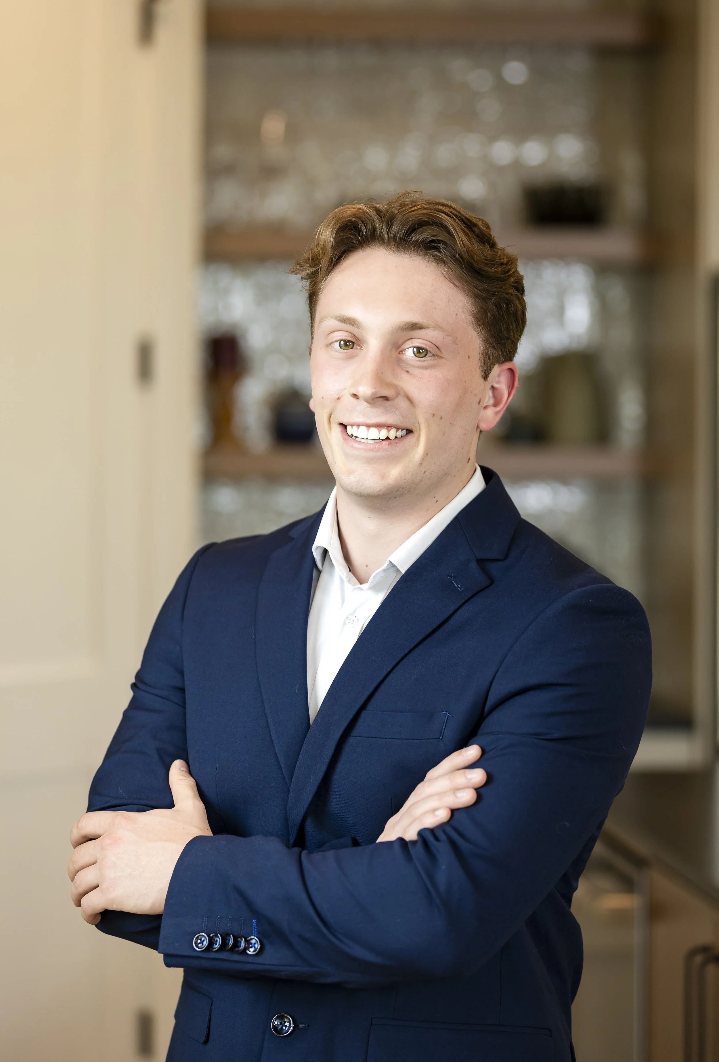 A young man with light skin, short brown hair, dressed in a navy suit and white shirt, posing with arms crossed in a kitchen setting.
